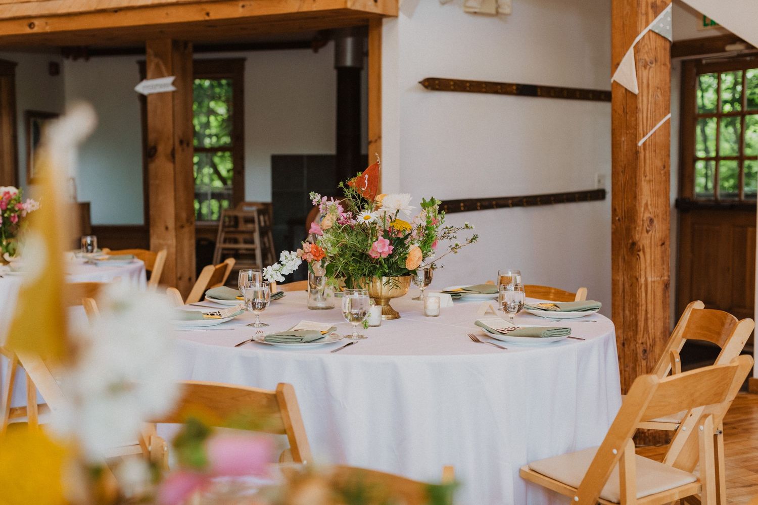 Elegant rustic wedding reception venue with round tables, wooden chairs, and colorful floral centerpieces in barn setting.