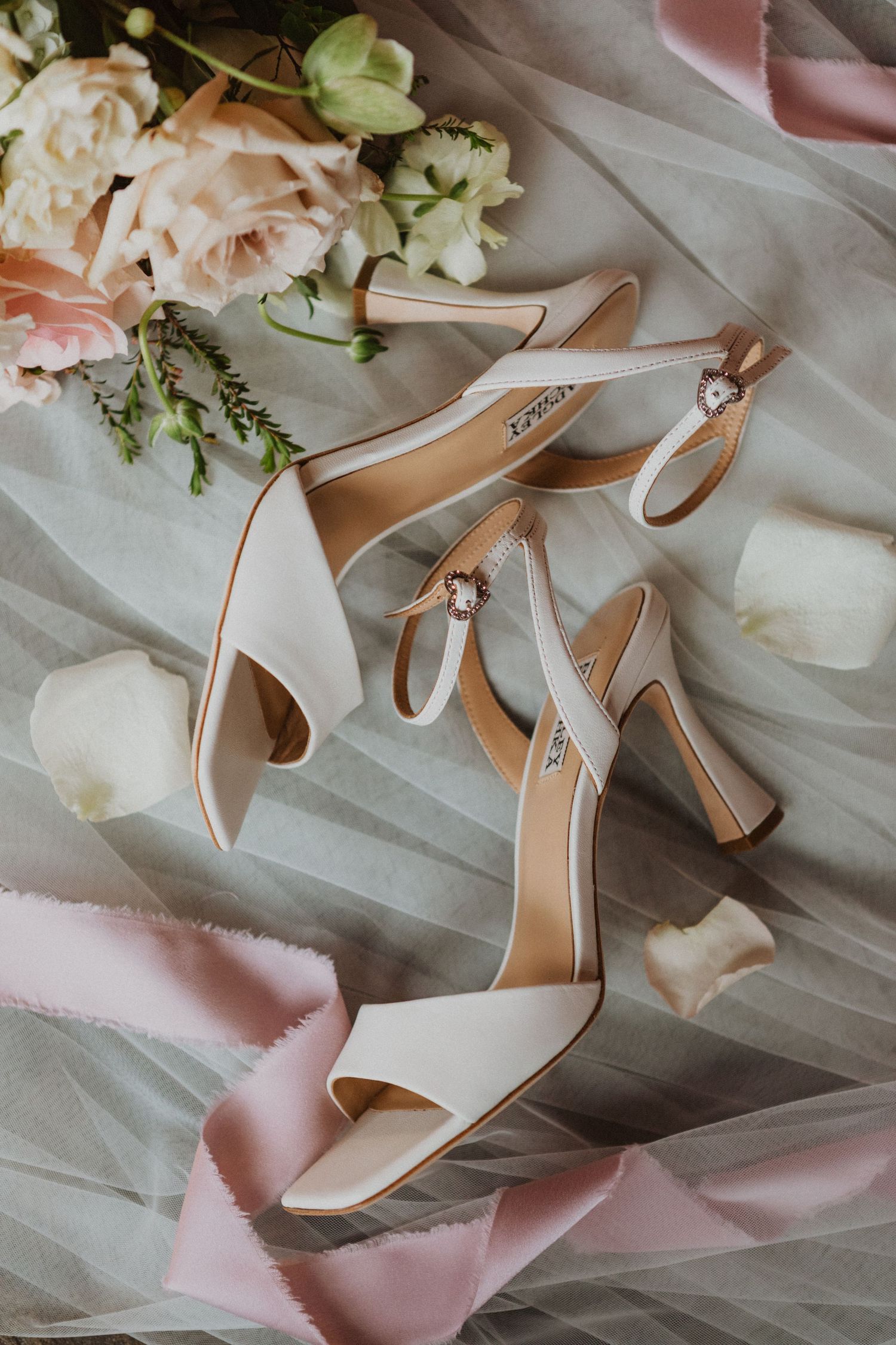 Elegant white block heel bridal sandals with ankle straps beside pink roses on sheer fabric.