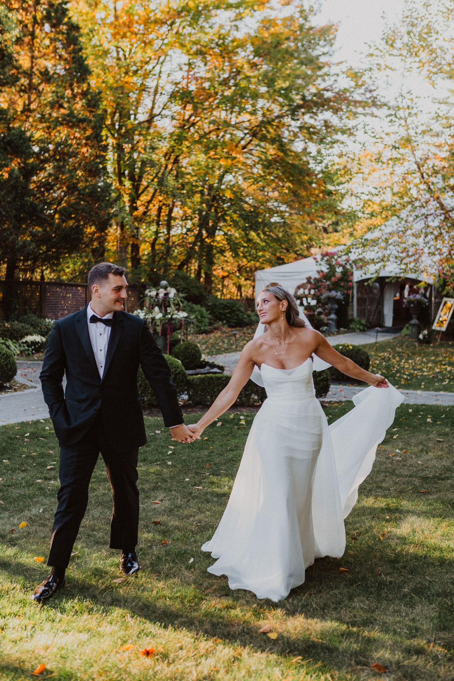Bride in white gown and groom in tuxedo walking hand in hand through autumn garden during wedding portraits.