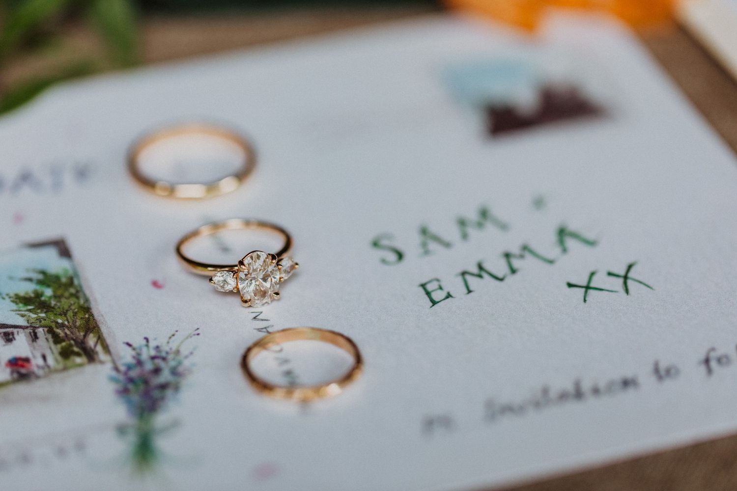 Gold wedding rings and diamond engagement ring displayed on a Sam and Emma wedding invitation with floral illustrations.