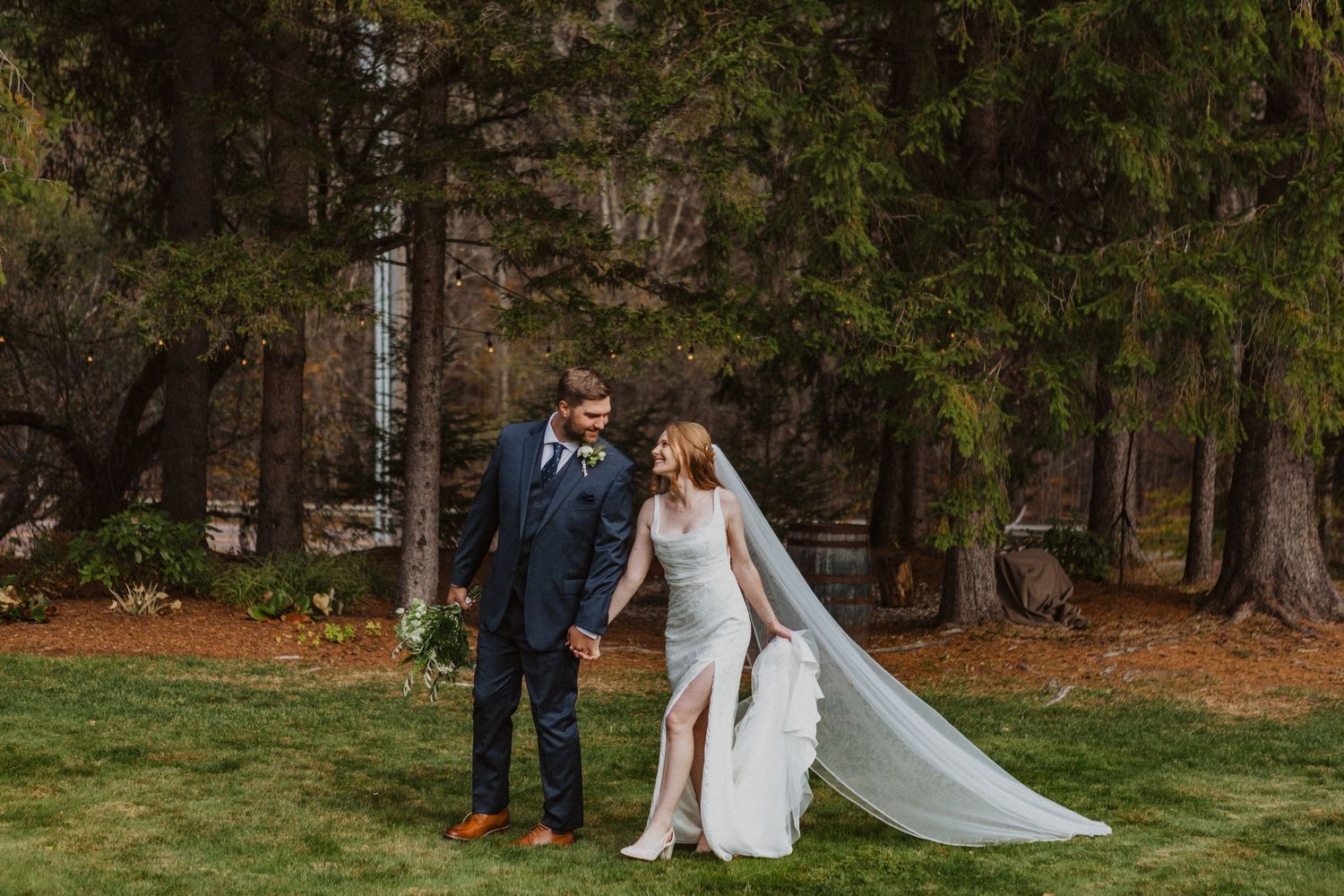 Bride and groom walking hand in hand through pine forest in elegant wedding attire.