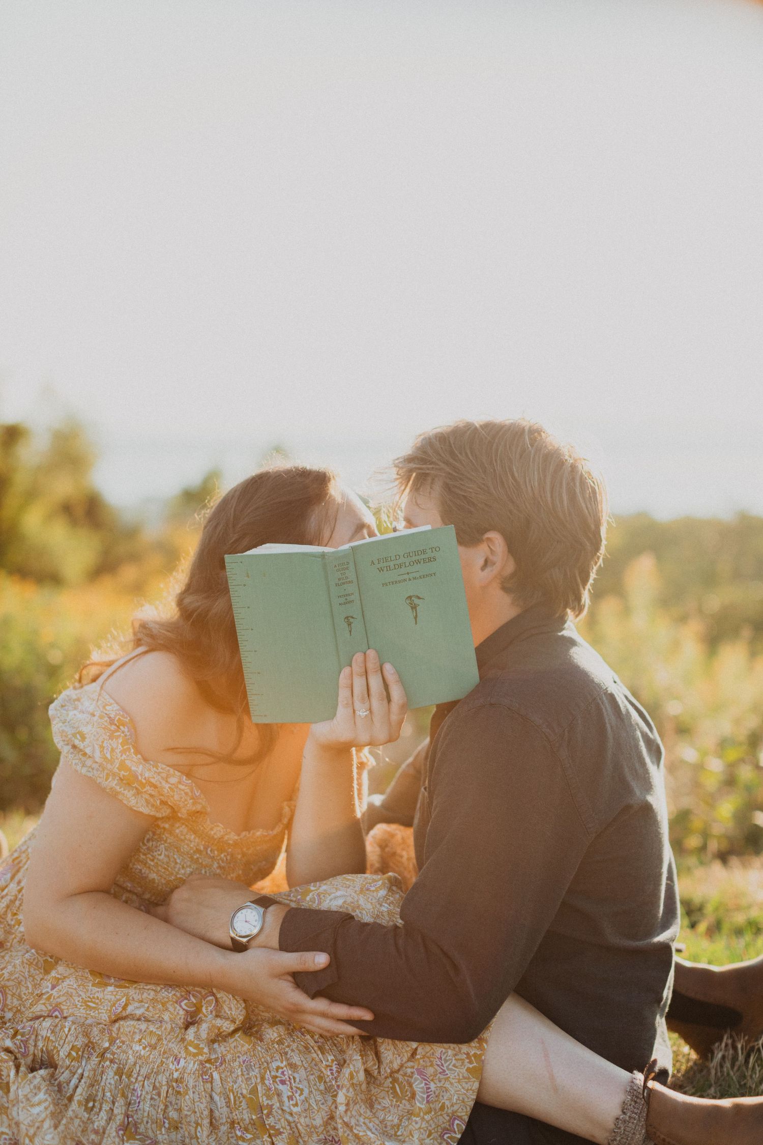 Couple kissing behind a teal book during romantic golden hour outdoor engagement photo session.