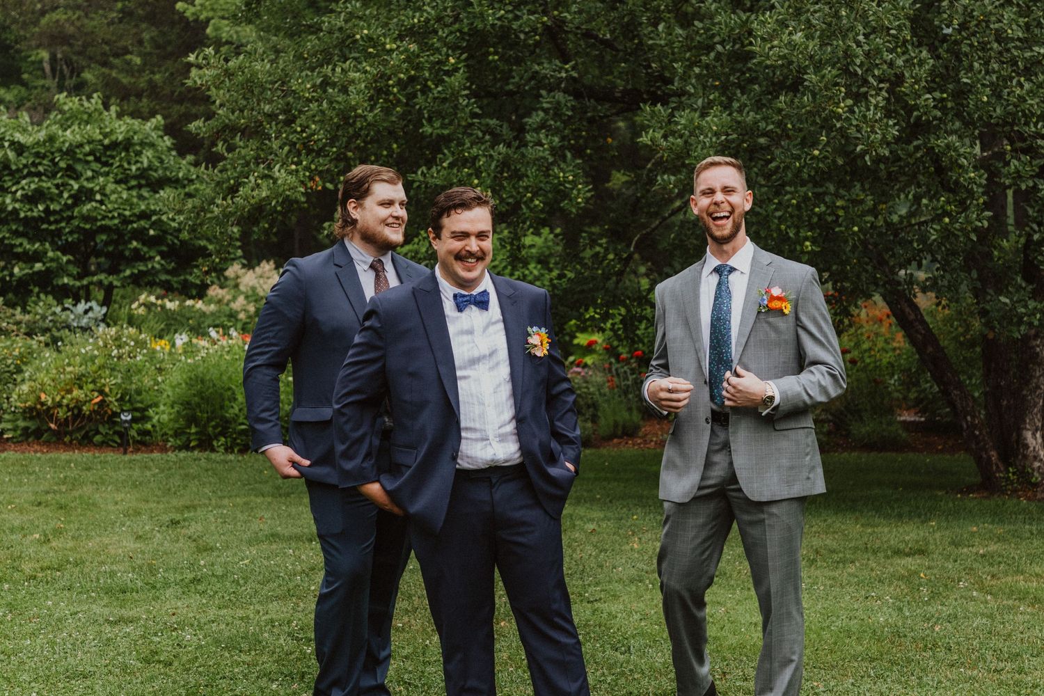 Three groomsmen in navy and grey suits laughing together in lush garden wedding venue.