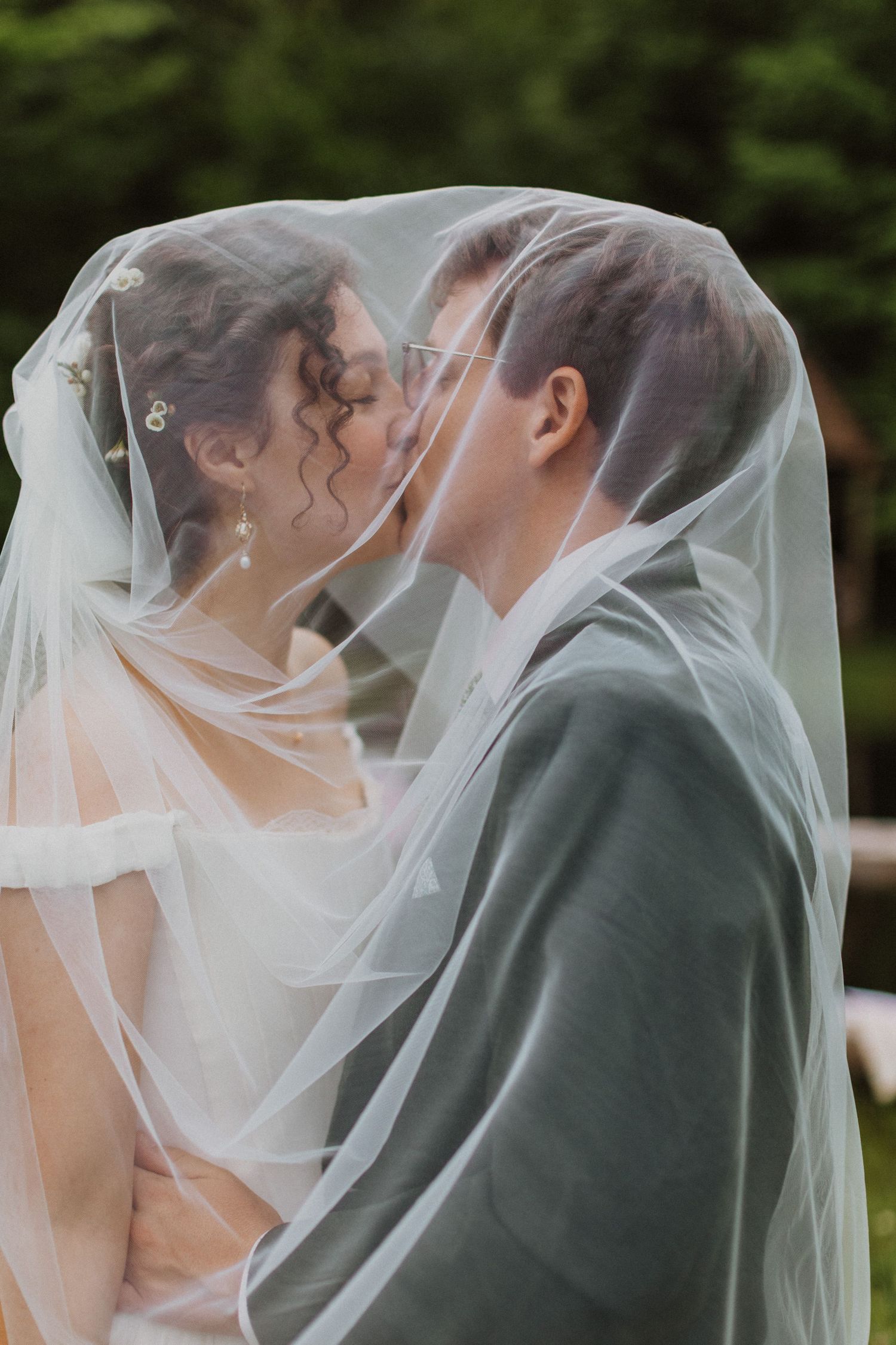 Romantic bride and groom sharing a kiss under sheer wedding veil outdoors in lush greenery.
