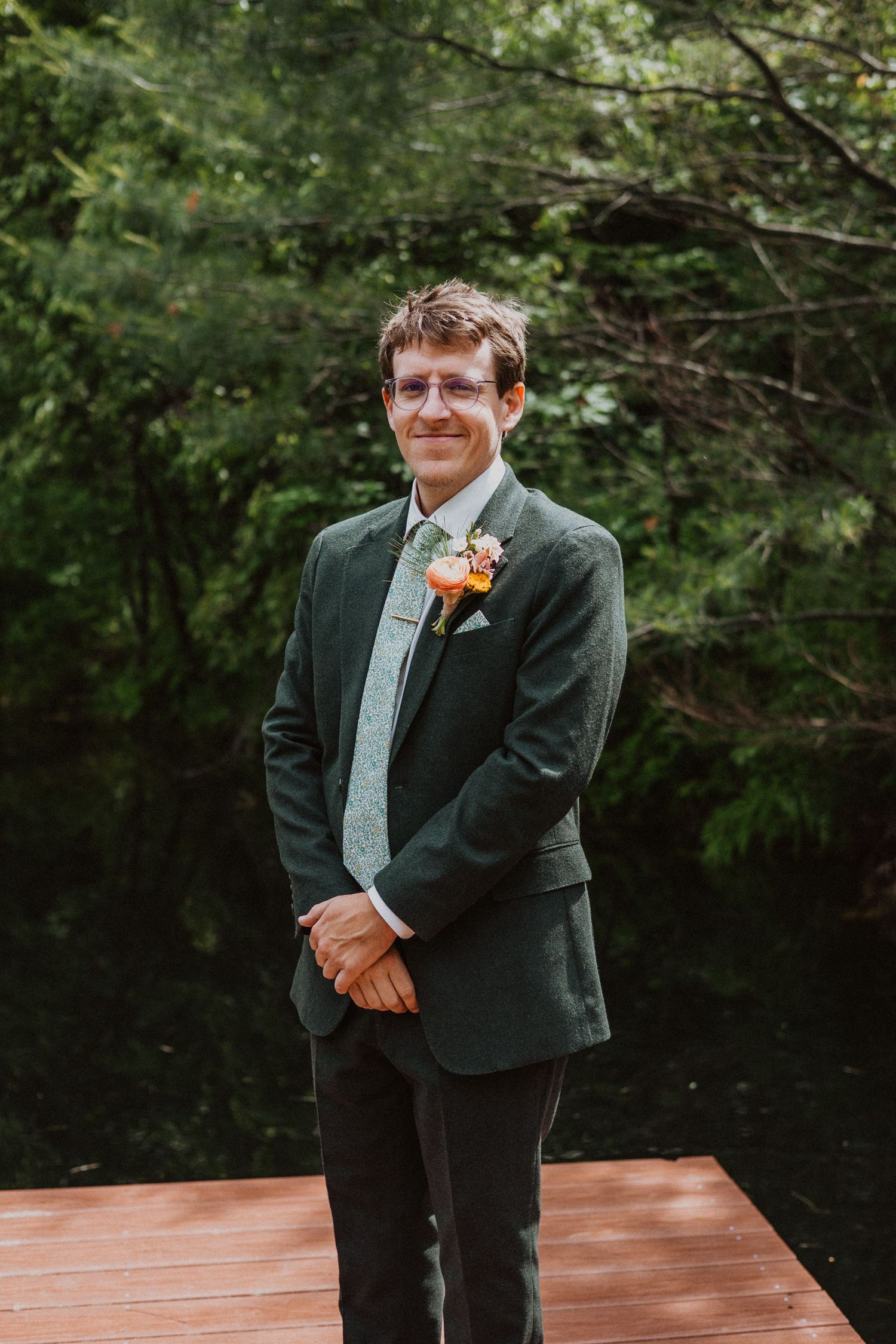 Groom in dark green suit with floral boutonniere standing on wooden deck surrounded by lush greenery.