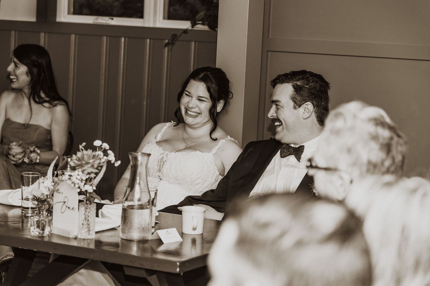 Bride and groom laughing together at wedding reception table in black and white photo.
