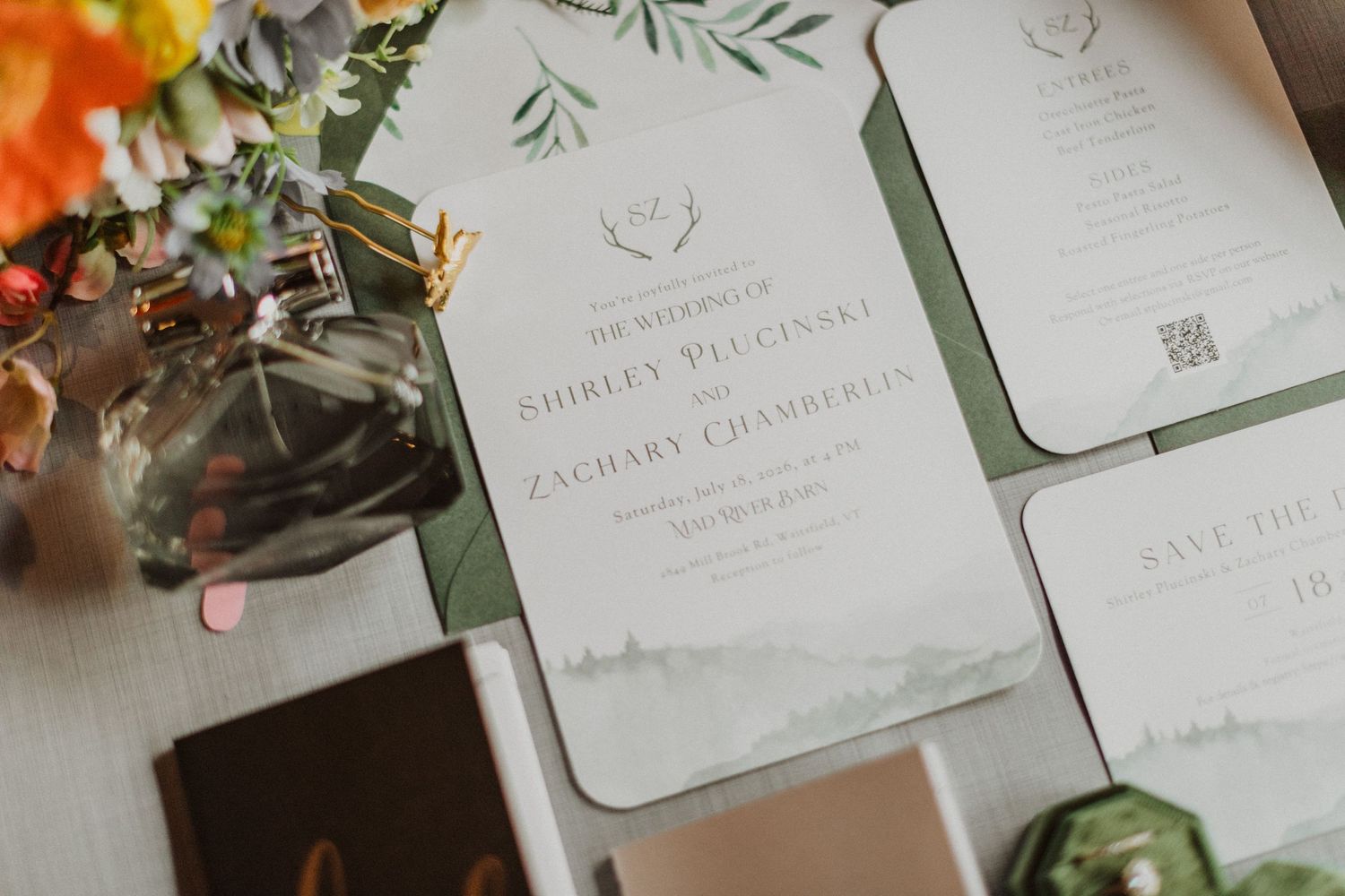 Elegant green and white wedding invitation suite with mountain watercolor design and floral accents displayed on a flat lay.