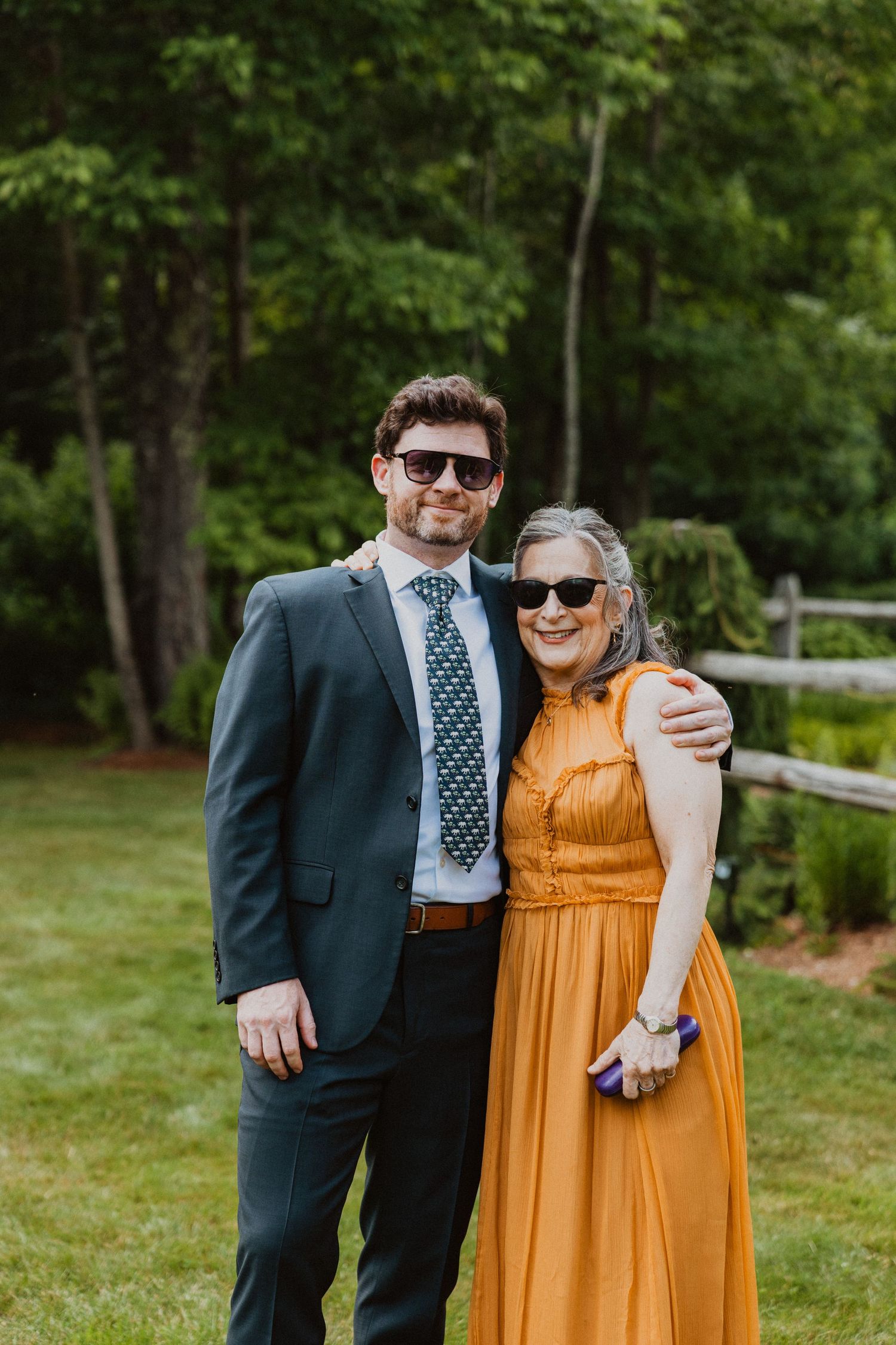 Groom in navy suit and older woman in gold dress wearing sunglasses pose together at outdoor wedding.