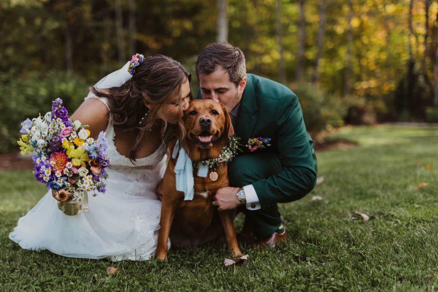 Bride and groom in green jacket kissing their brown dog wearing a bandana at outdoor wedding.