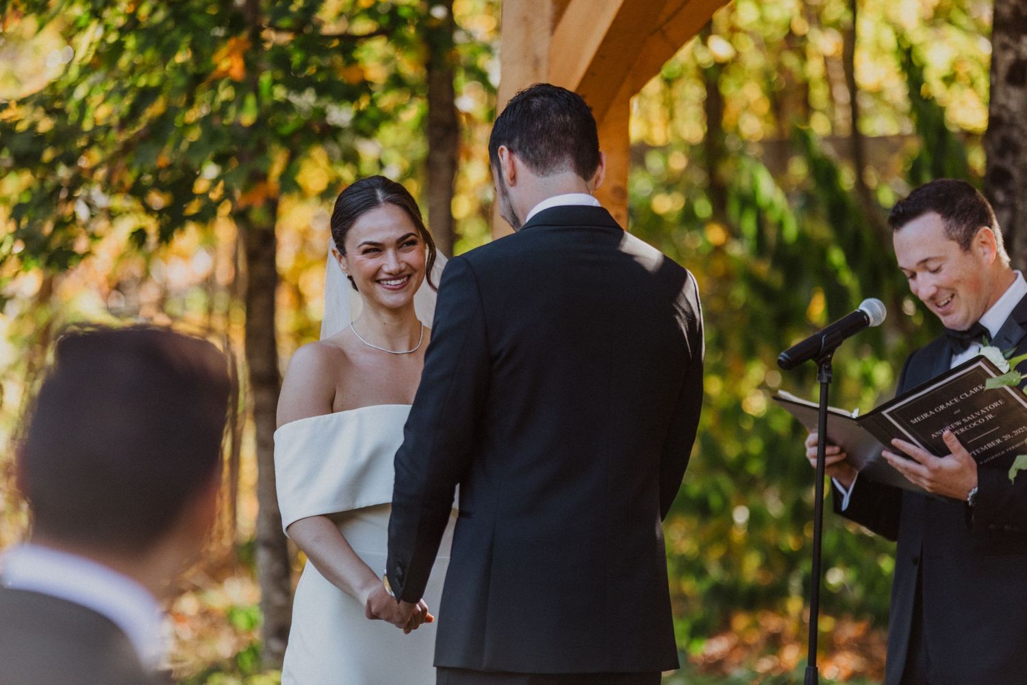 Radiant bride laughing while holding grooms hands during outdoor autumn wedding ceremony.