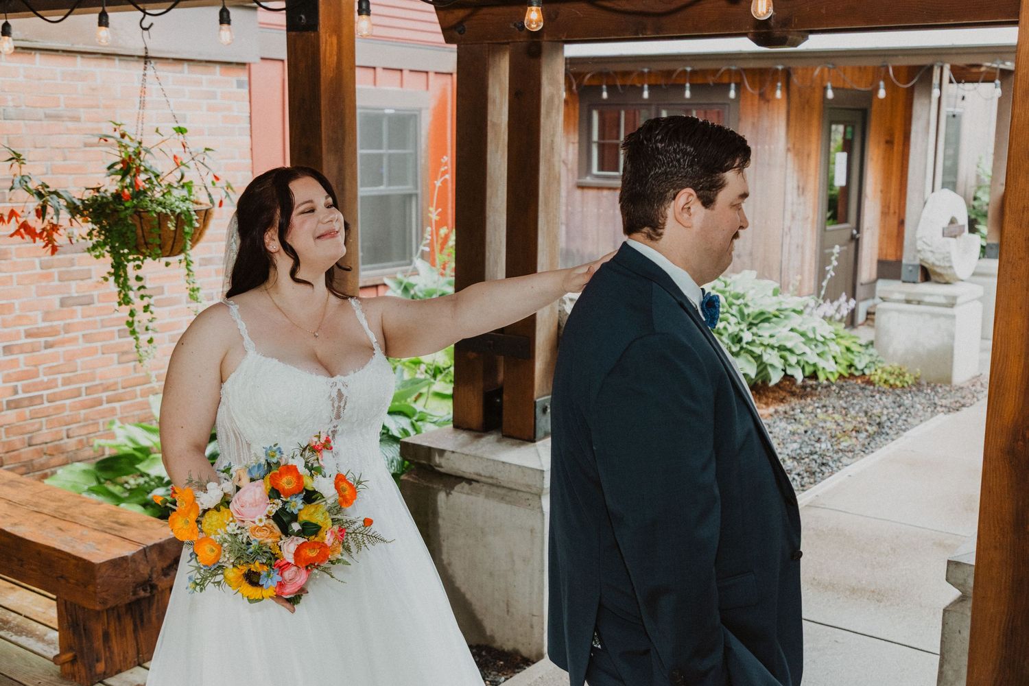Bride in white gown holding colorful bouquet taps groom's shoulder during first look at rustic outdoor wedding venue.