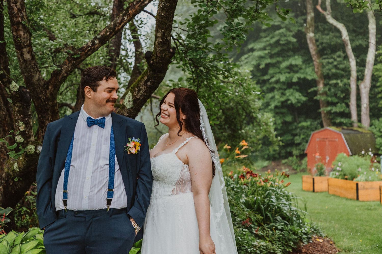 A bride and groom share a joyful moment outdoors, surrounded by lush greenery, with a red barn visible in the background.