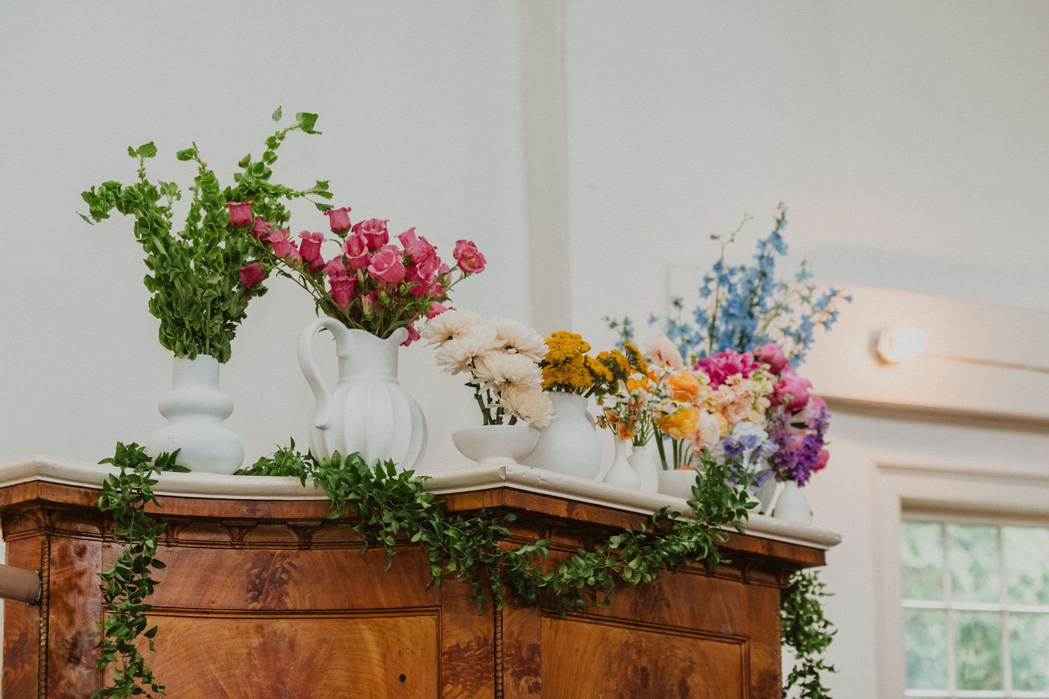 Colorful floral arrangements in white pitchers and vases on wooden dresser with green garland.