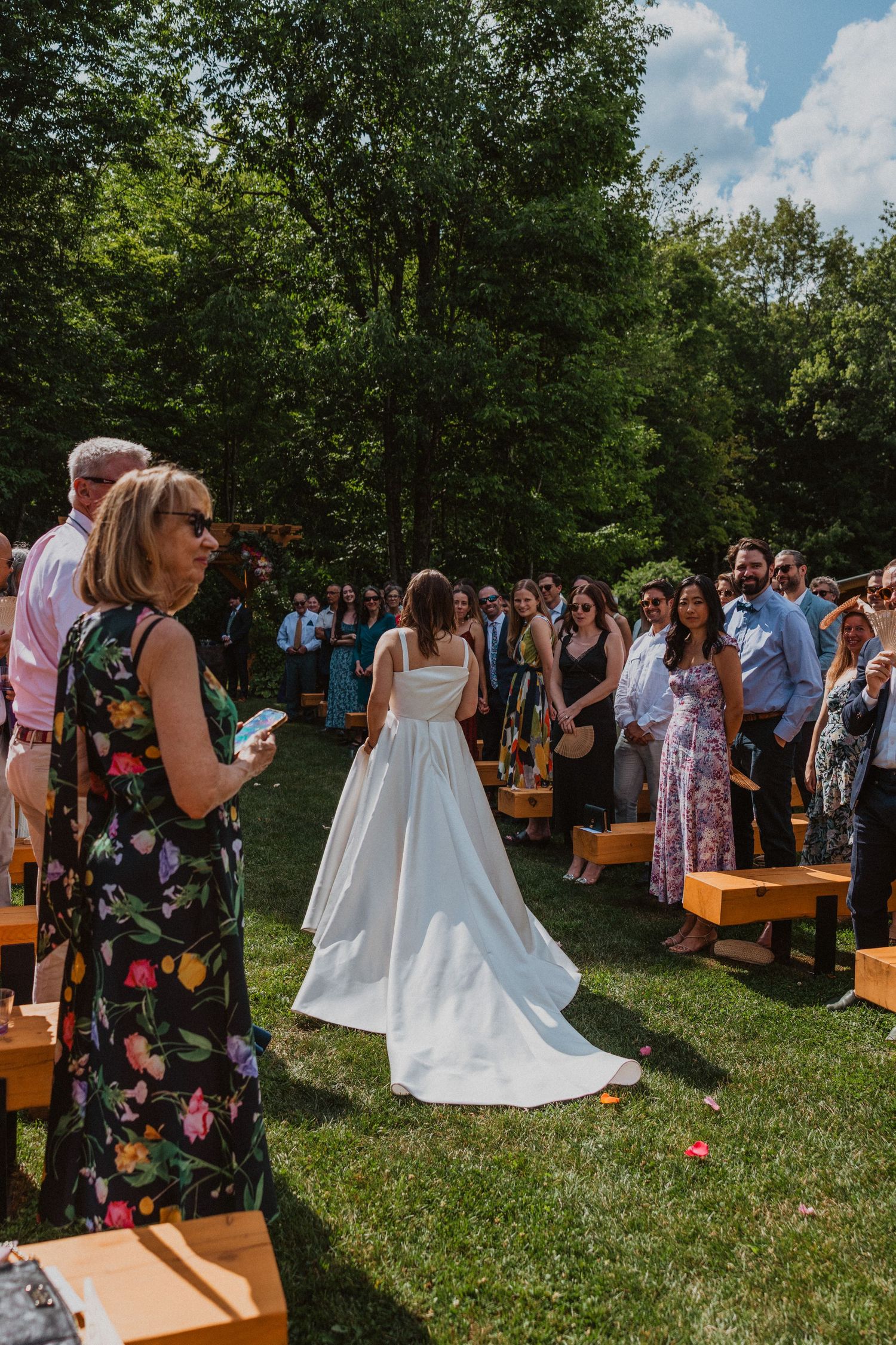 Bride walks down outdoor aisle in white gown as wedding guests watch ceremony.