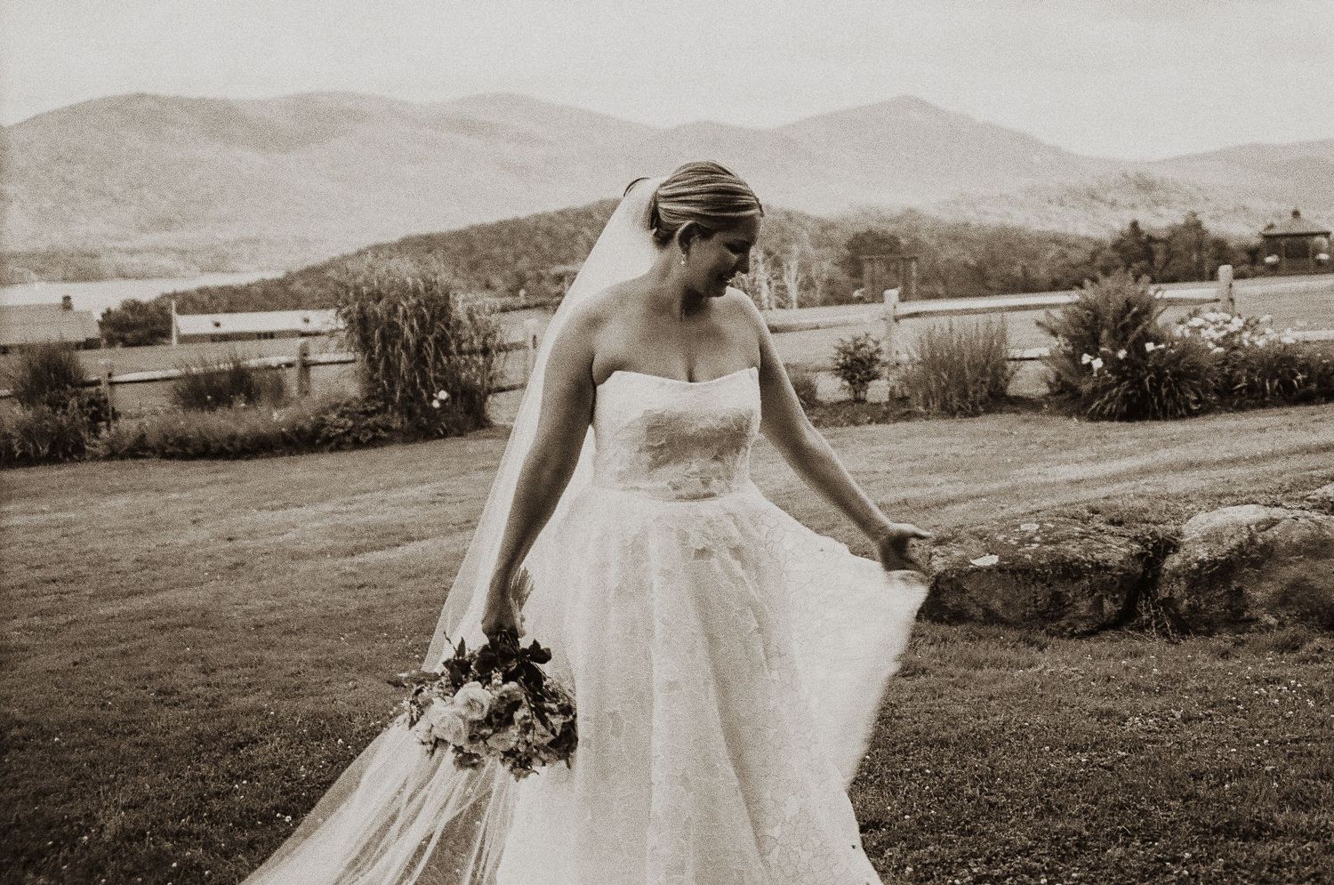 Sepia-toned bride in strapless gown holding bouquet with mountain landscape in background.
