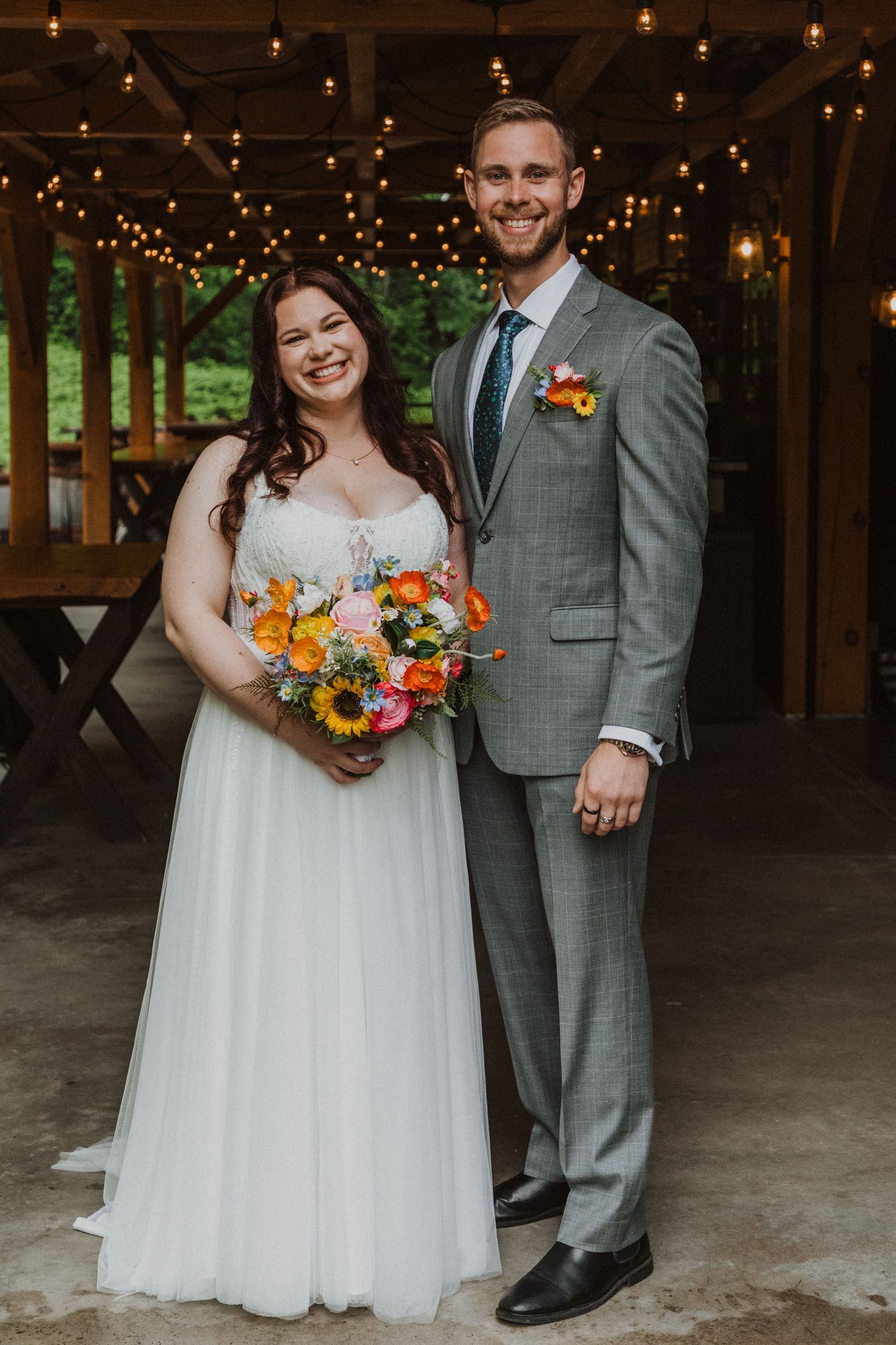 Bride in white gown holding colorful sunflower bouquet poses with groom in gray suit under string lights at rustic venue.
