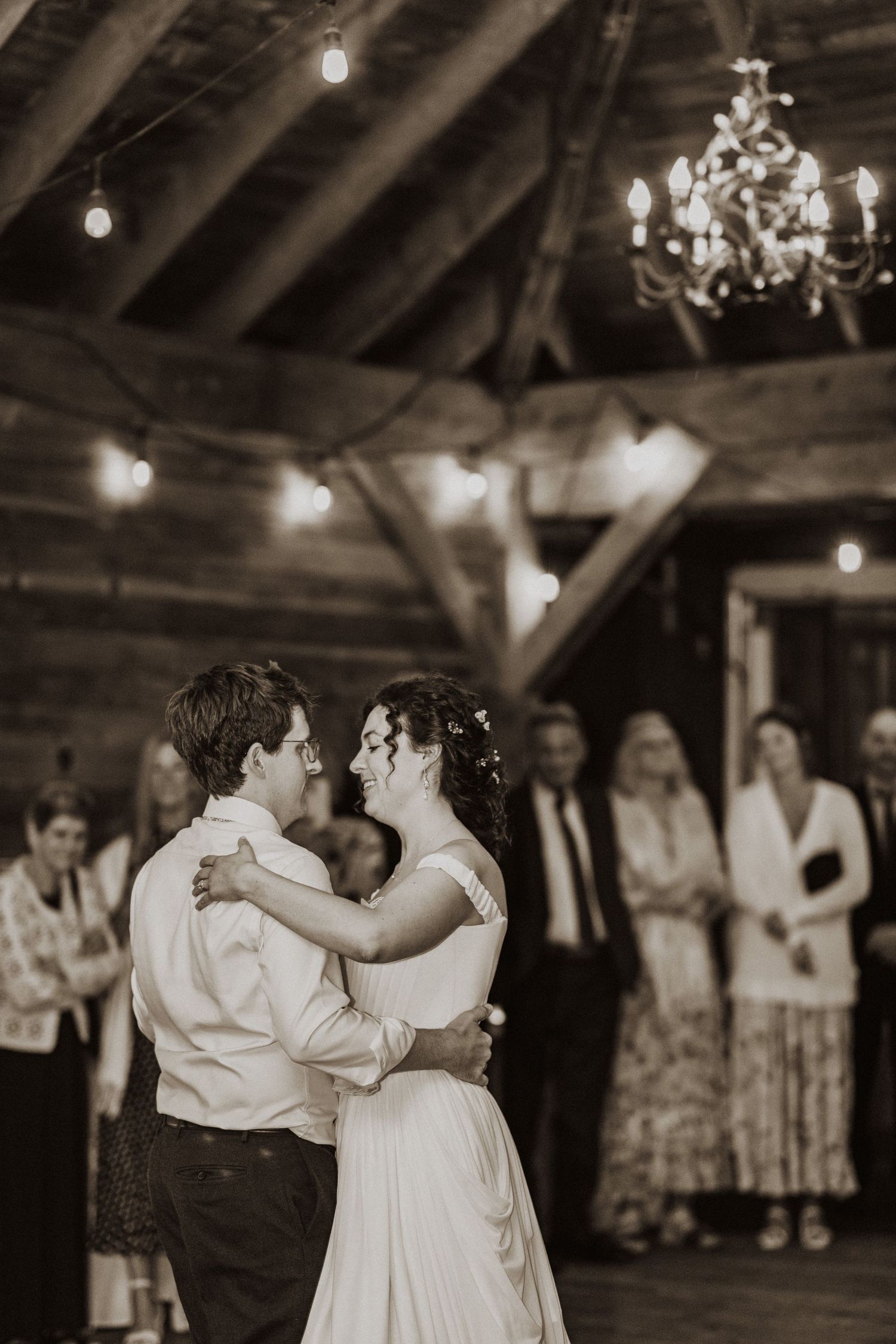 Sepia-toned first dance photo of newlyweds in a rustic barn with string lights and chandelier.