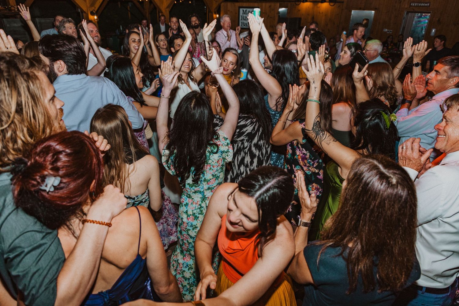 Energetic wedding reception dance floor with guests raising hands and dancing joyfully in colorful outfits.