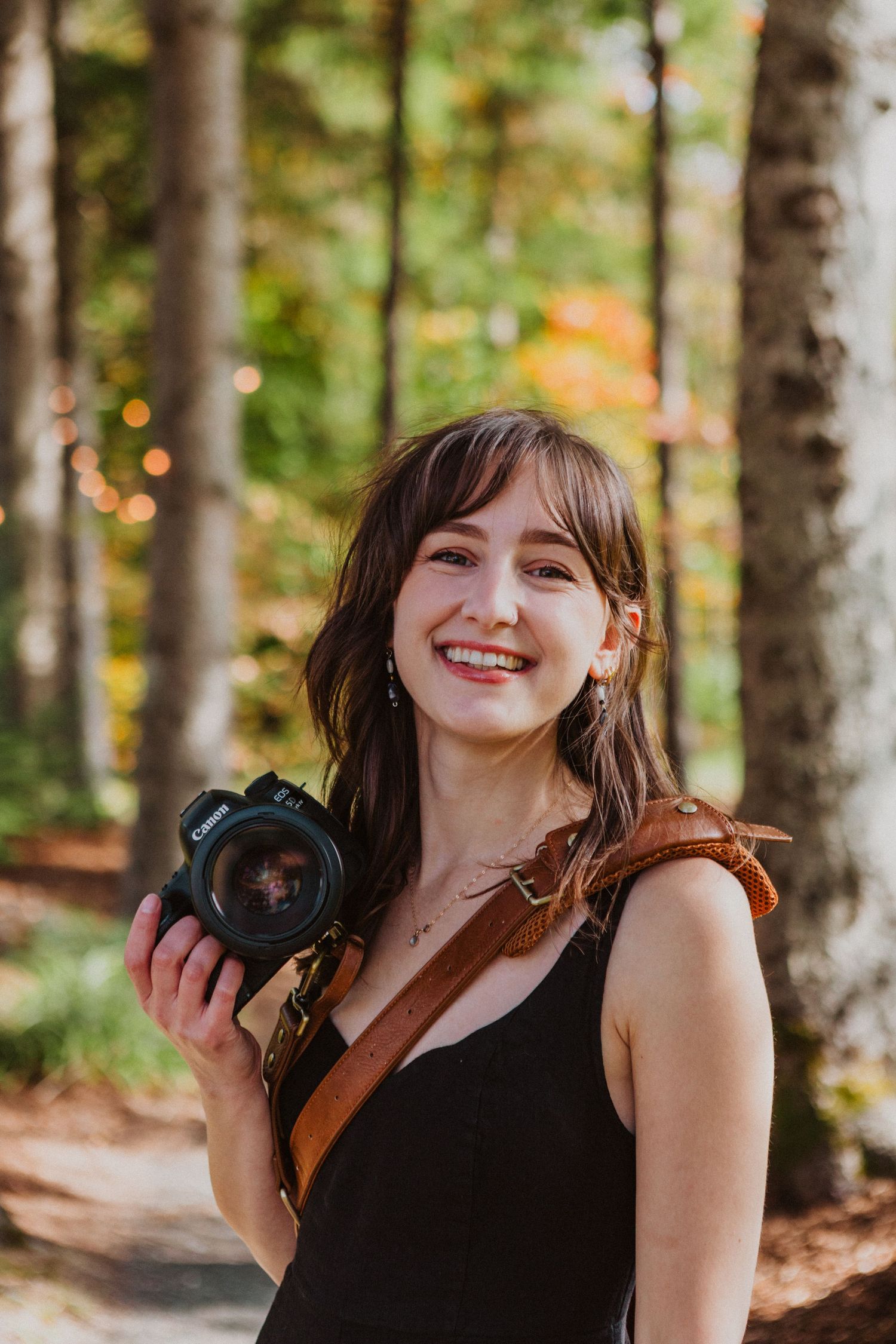 Smiling female photographer holds camera in autumn forest with colorful foliage background.