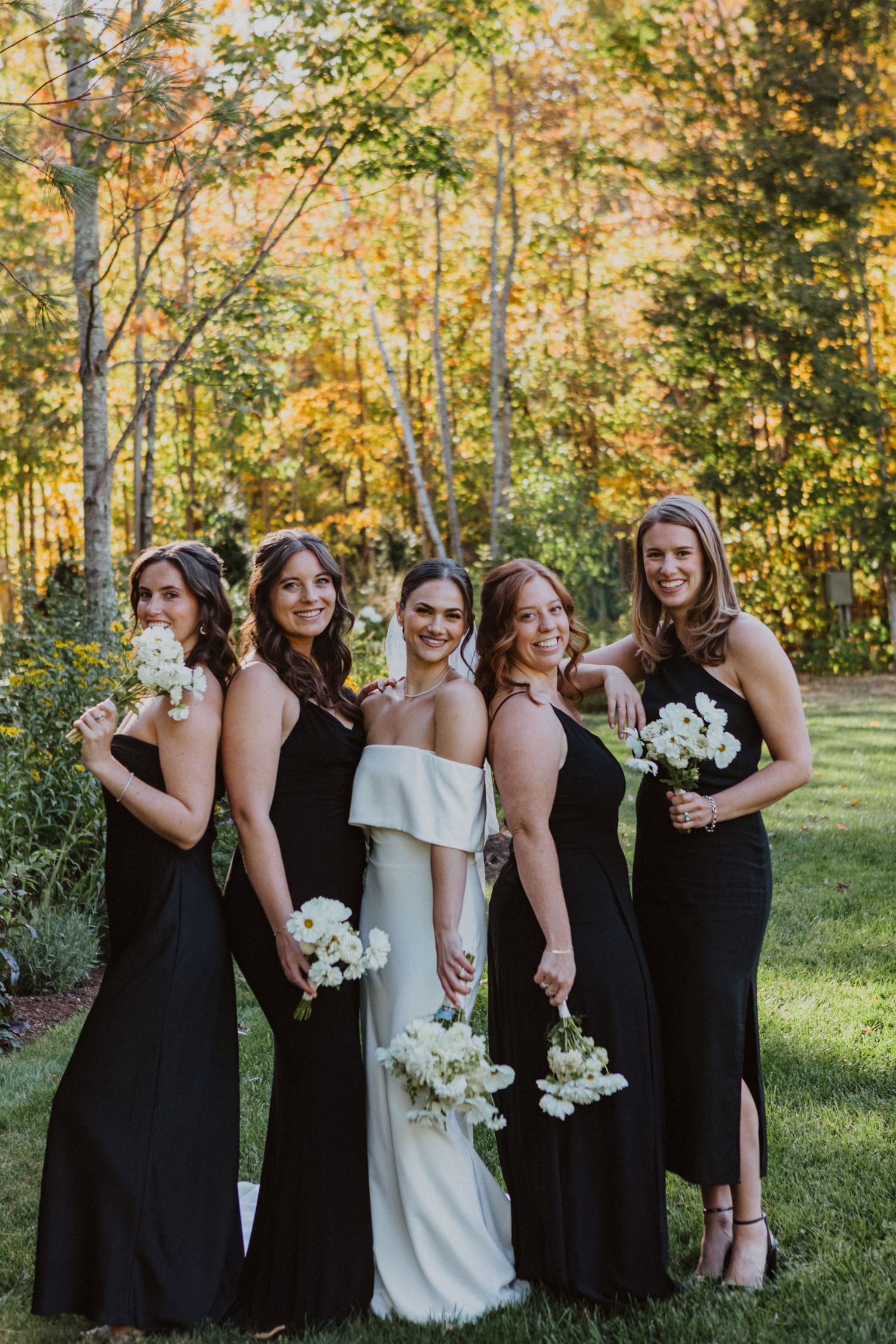 Bride in white off-shoulder gown poses with four bridesmaids in black dresses holding white bouquets outdoors.