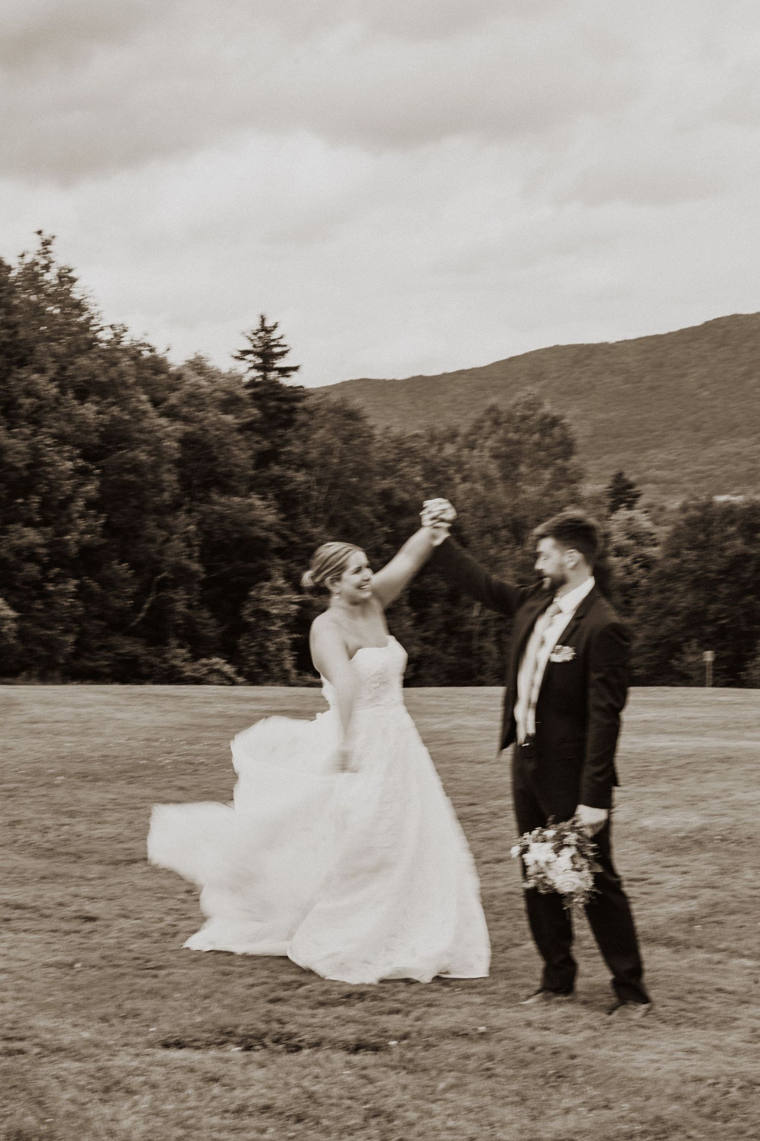 Black and white photo of groom twirling bride in flowing gown on a scenic mountain field.