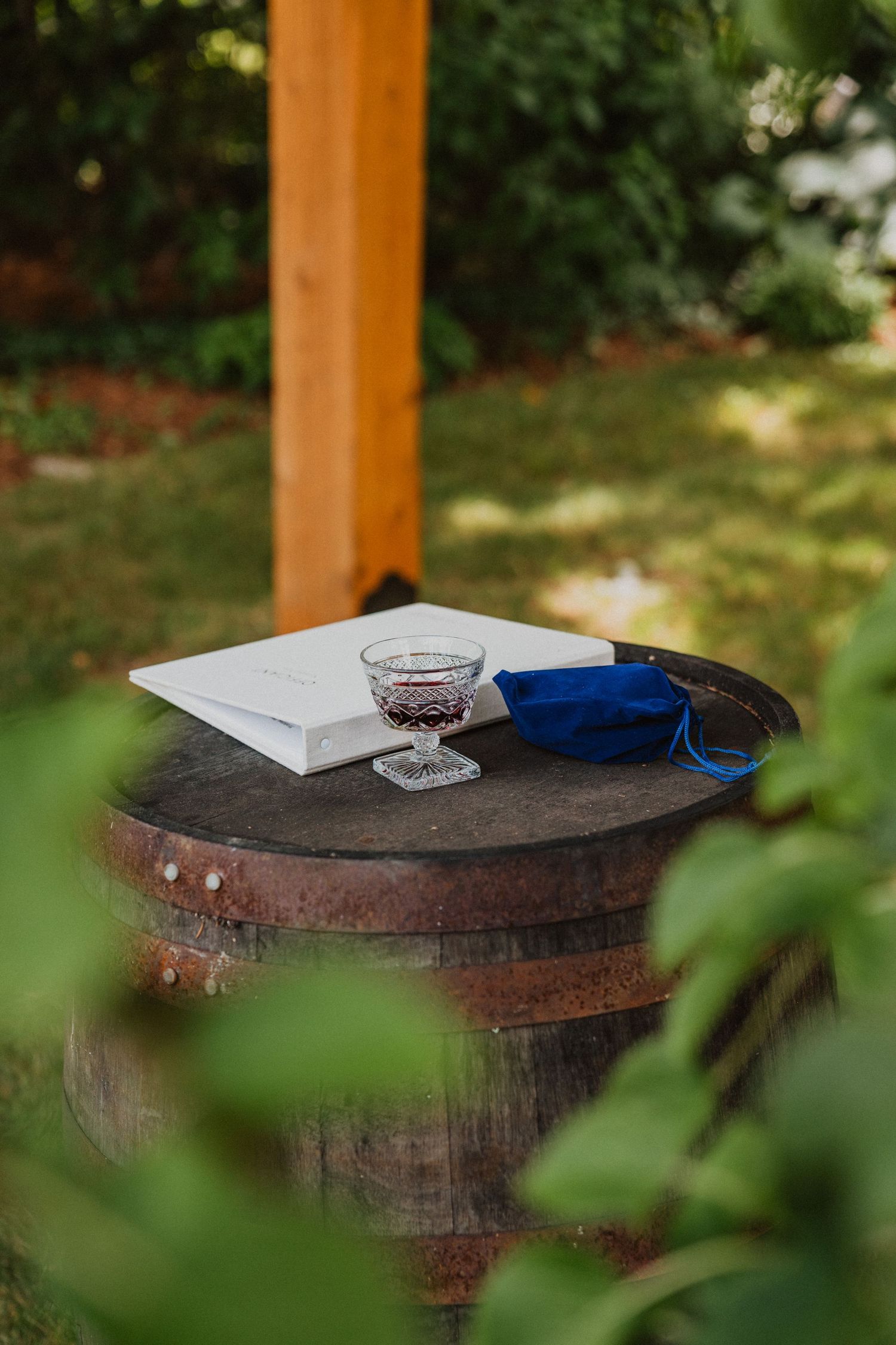 A rustic wooden barrel topped with a white book, crystal glass, and blue fabric at an outdoor wedding ceremony.