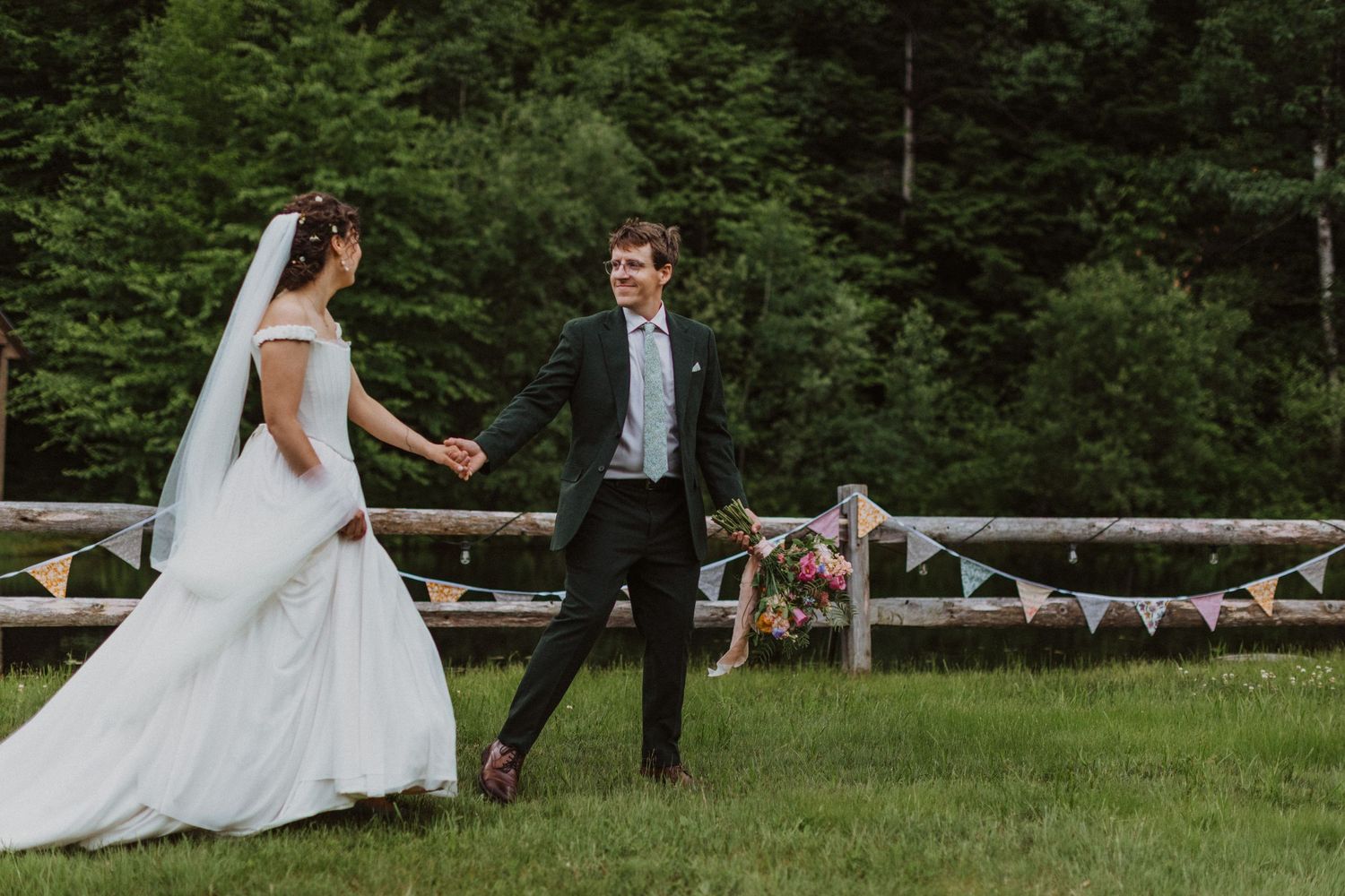 Bride and groom walking hand in hand on green lawn with rustic fence and bunting at outdoor wedding.
