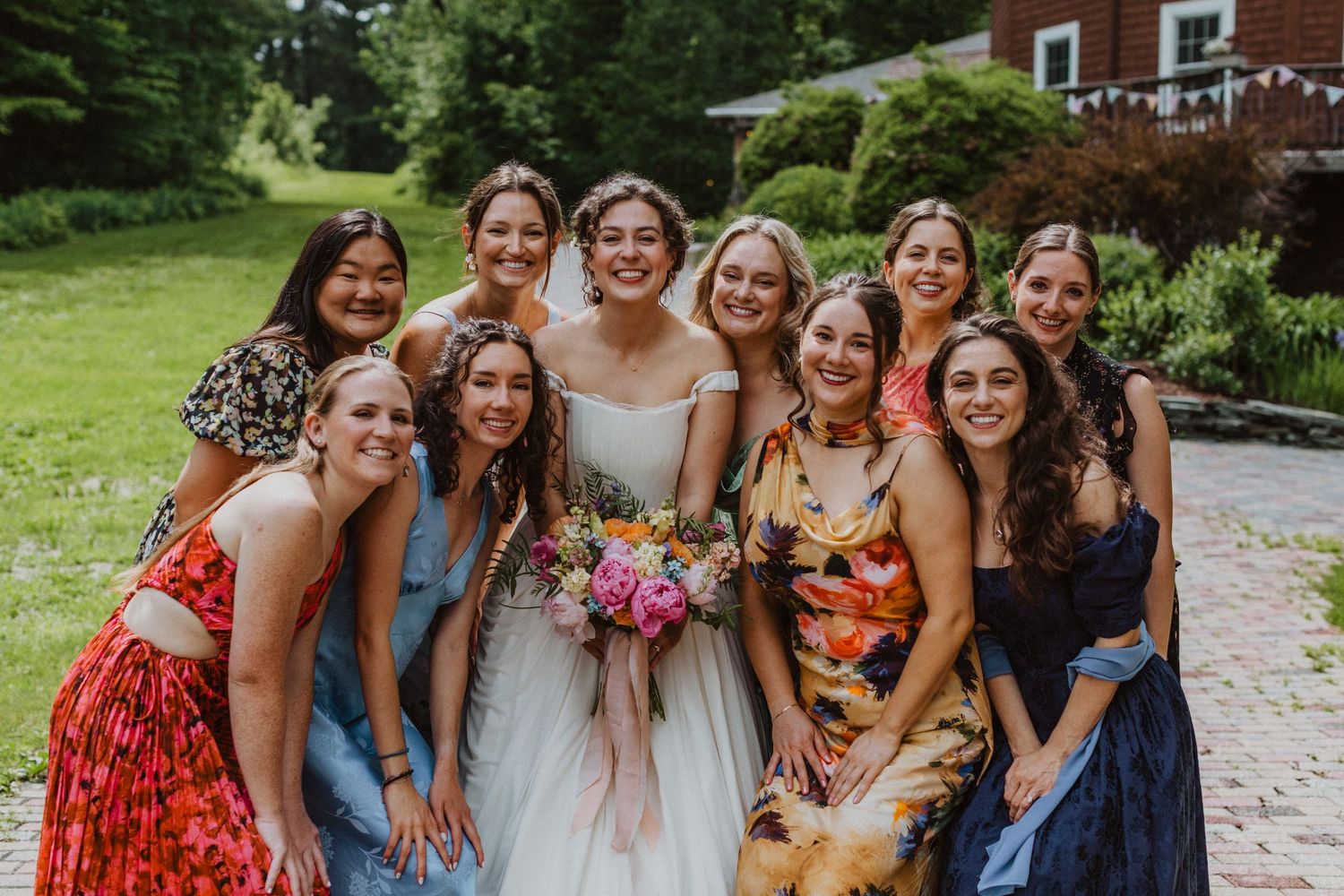Bride in white gown holds colorful bouquet surrounded by smiling bridesmaids outdoors.