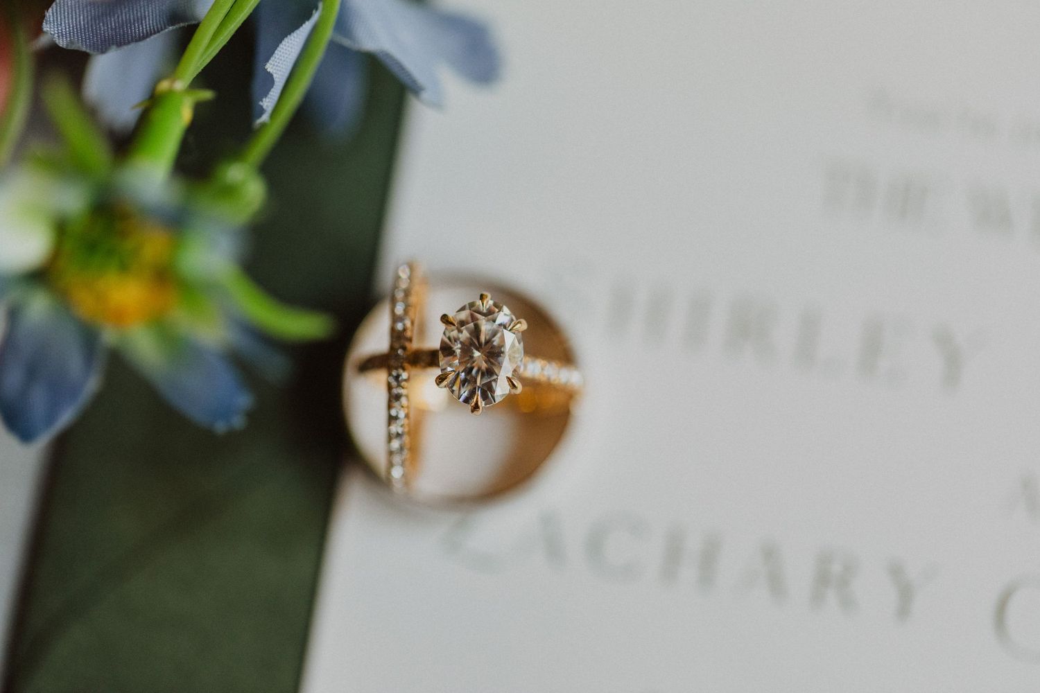Gold engagement ring with diamond solitaire and cross band displayed on wedding invitation for Shirley and Zachary.