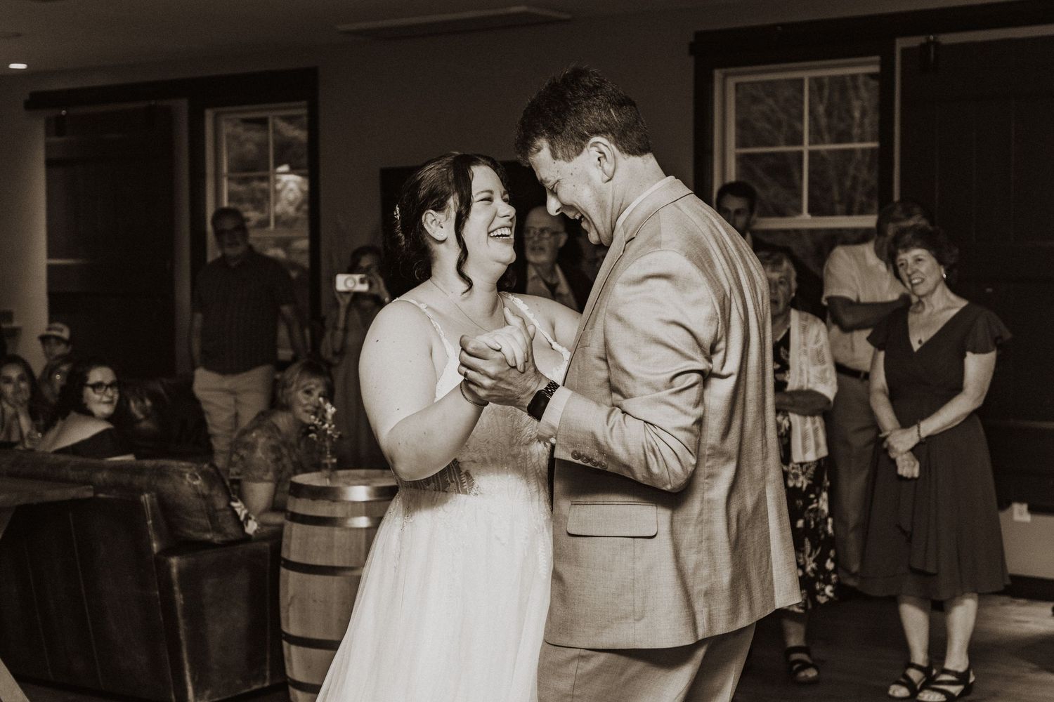 Bride and groom share first dance at rustic barn wedding reception in warm sepia-toned photograph.