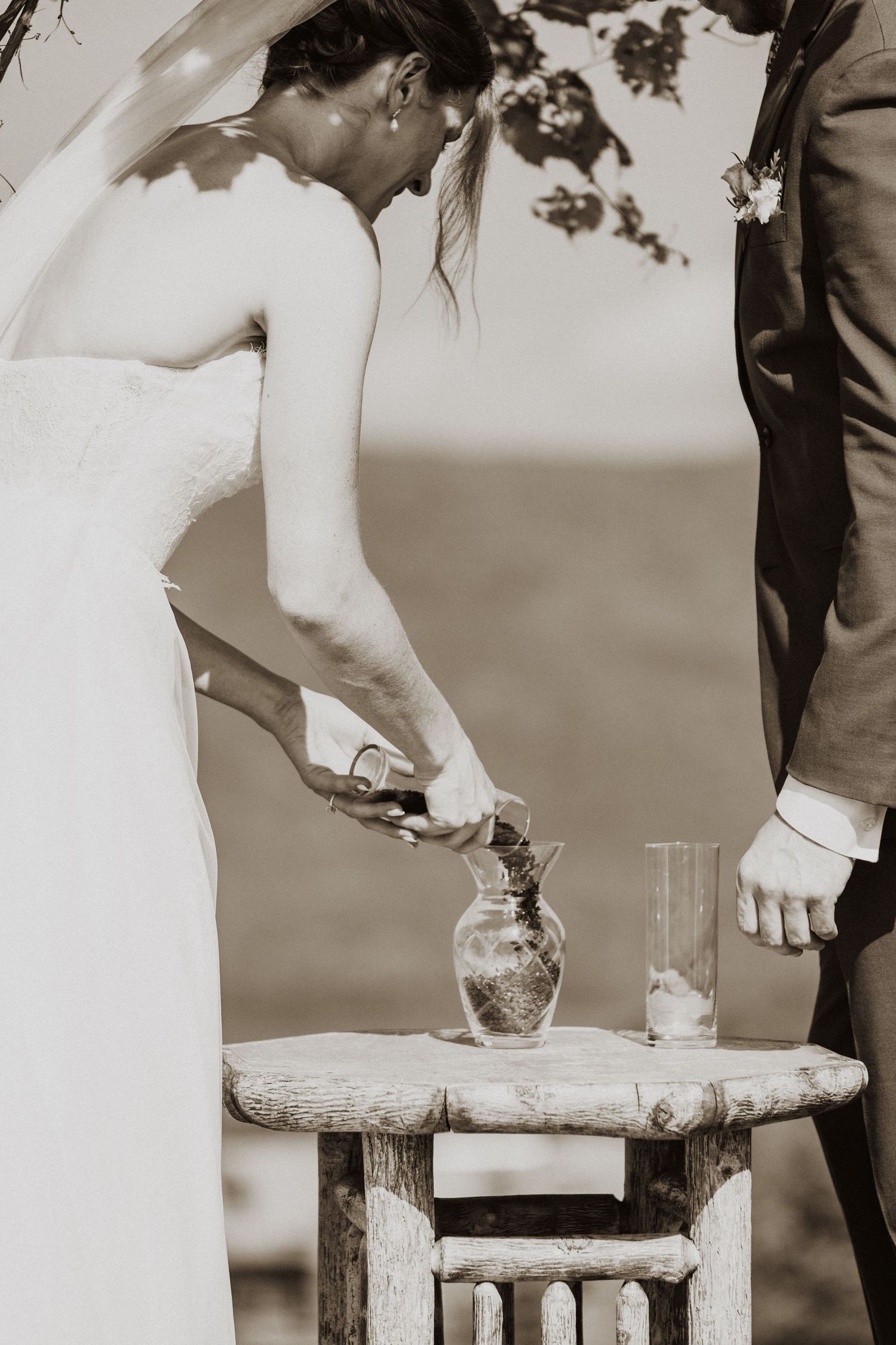 Bride and groom performing sand ceremony at rustic wooden table during outdoor wedding.