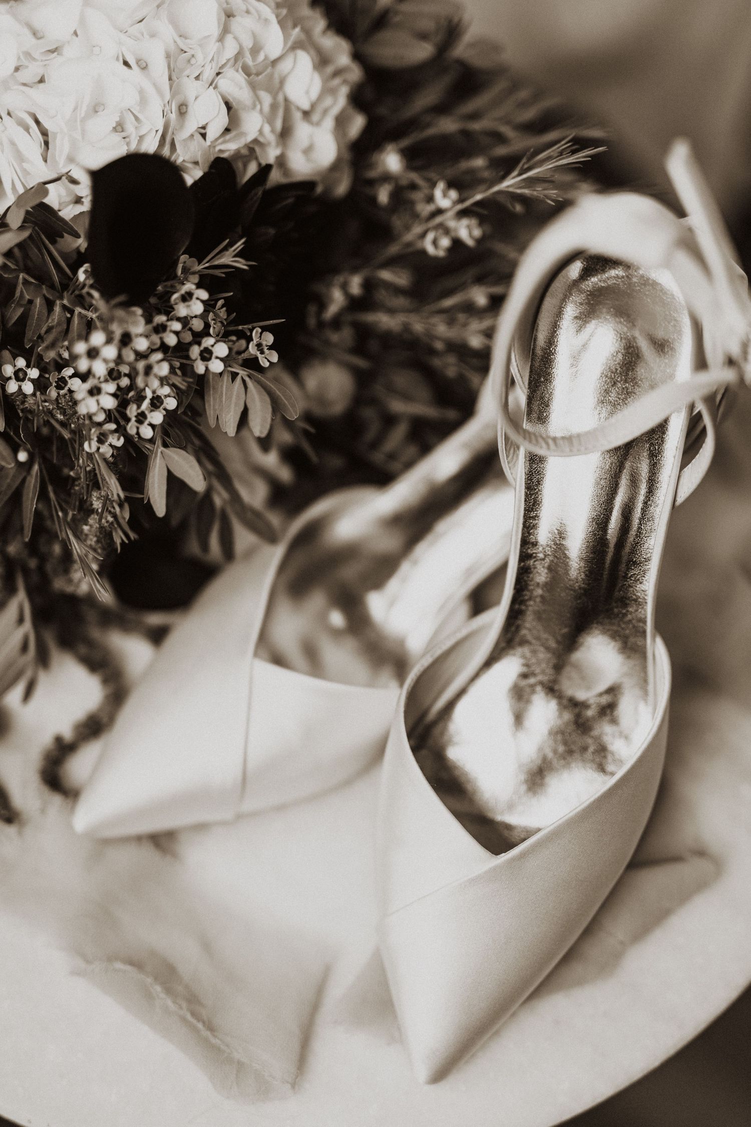 Black and white close-up of elegant white bridal heels beside a floral bouquet with greenery.