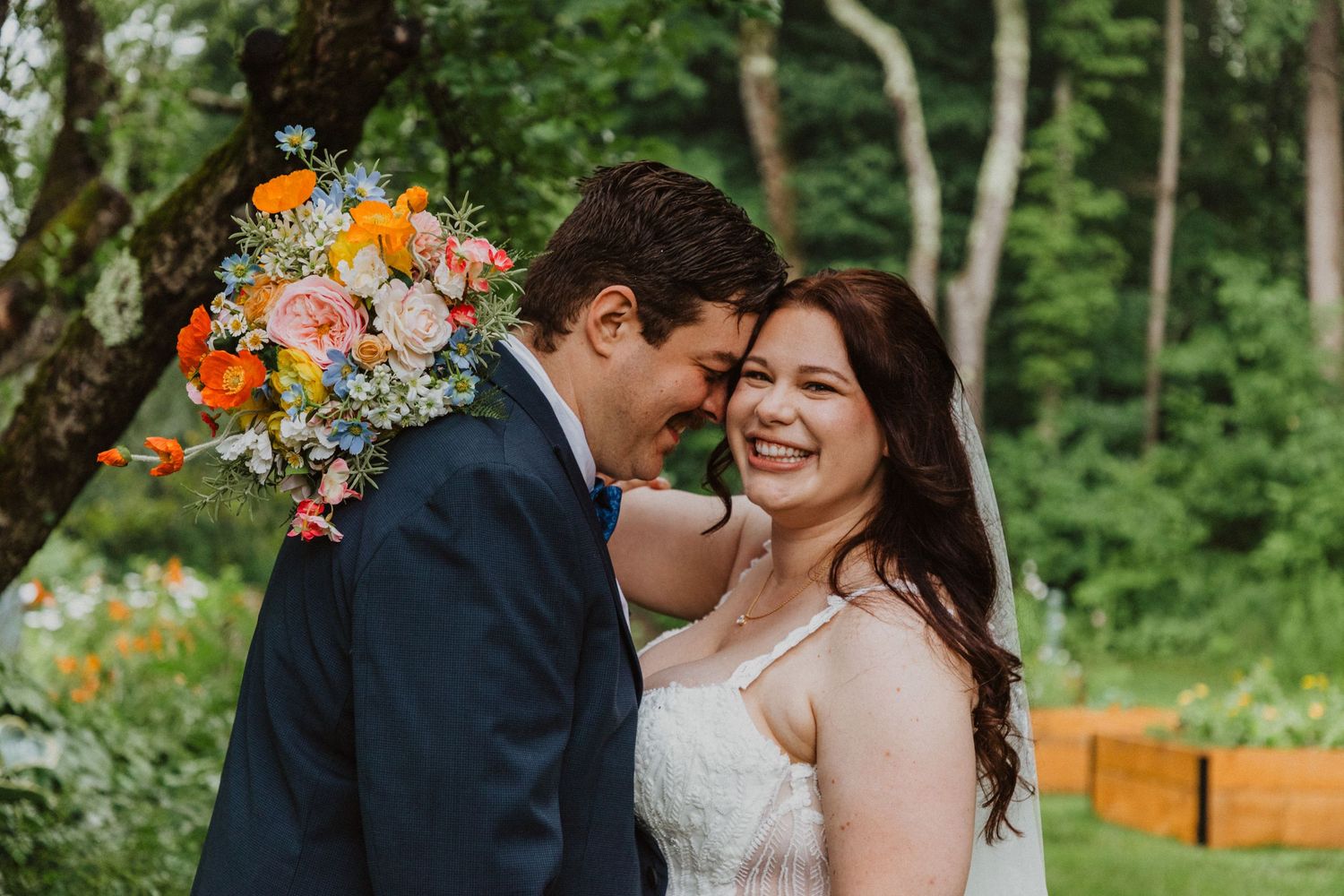 A joyful bride and groom share a tender moment outdoors, with a vibrant orange and pink bridal bouquet in a lush garden setting.