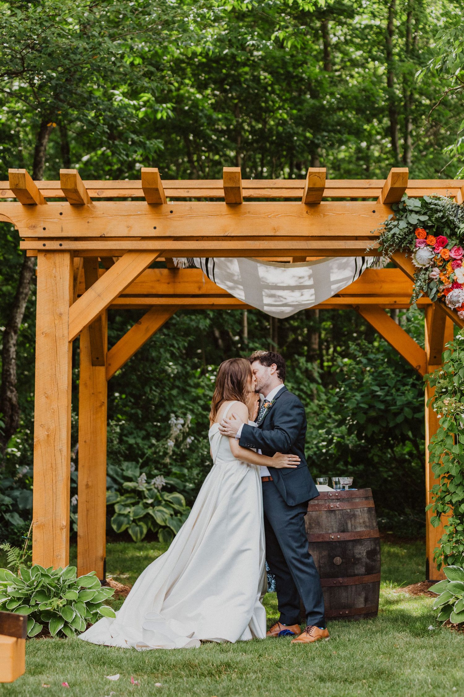 Bride and groom share first kiss under wooden pergola with floral decor at outdoor garden wedding ceremony.