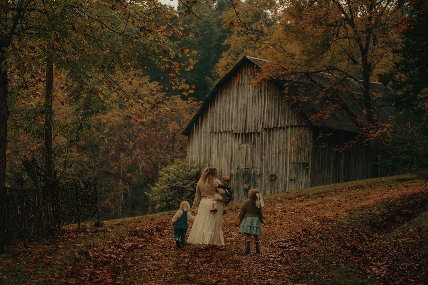 Bride walks with two children toward rustic wooden barn surrounded by colorful autumn foliage in a forest setting.
