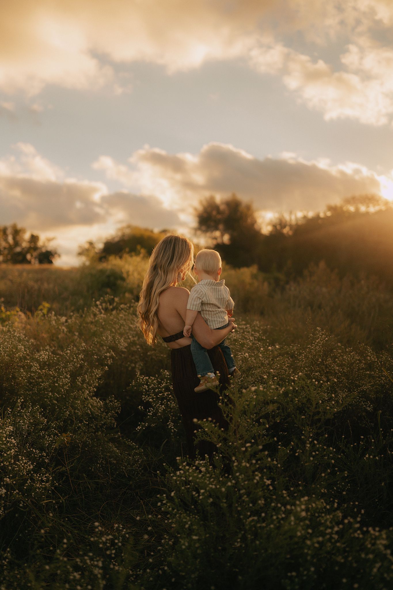 Mother holding baby in golden sunset field of wildflowers, warm light glowing through clouds.