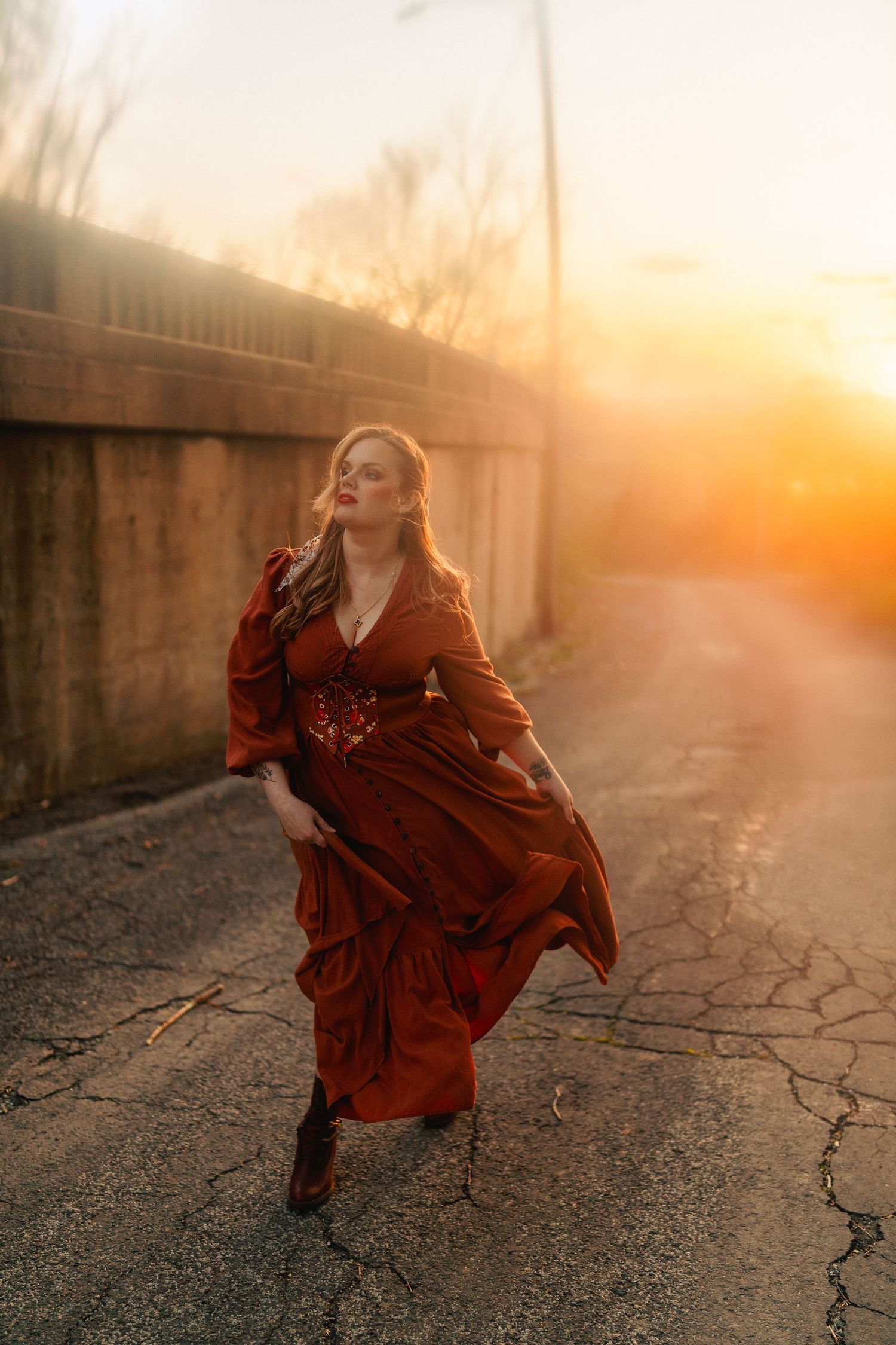 Woman in flowing rust-red dress walking on cracked road at golden sunset with dramatic light.