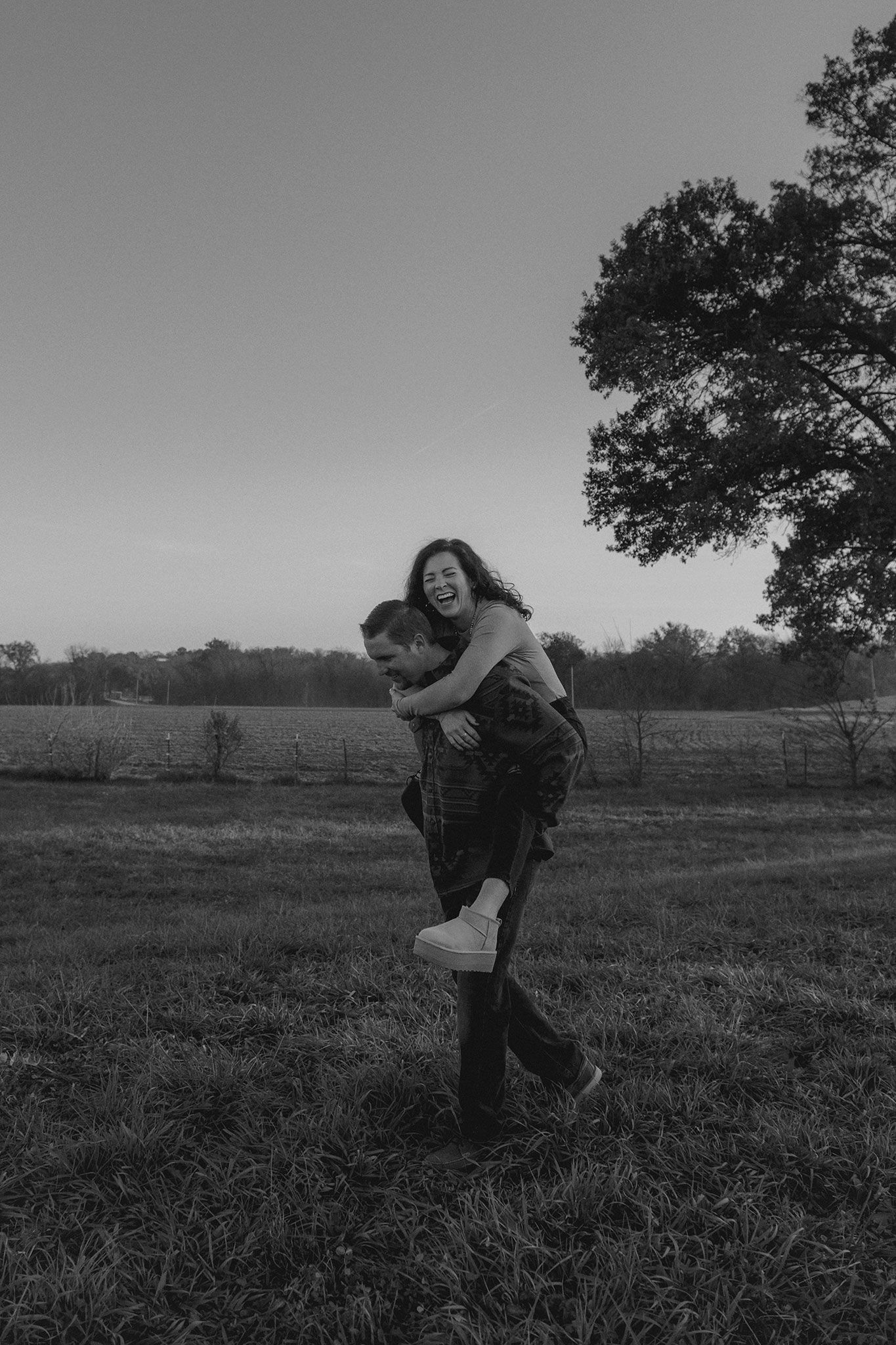 A man gives a laughing woman a piggyback ride across an open field at dusk in black and white.