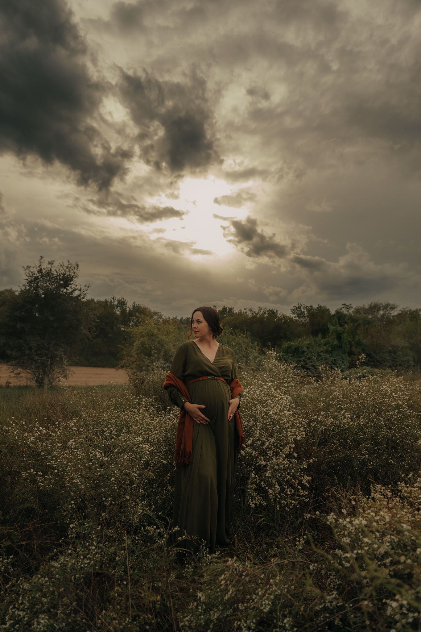Pregnant woman in green wrap dress standing in wildflower field under dramatic cloudy sky.