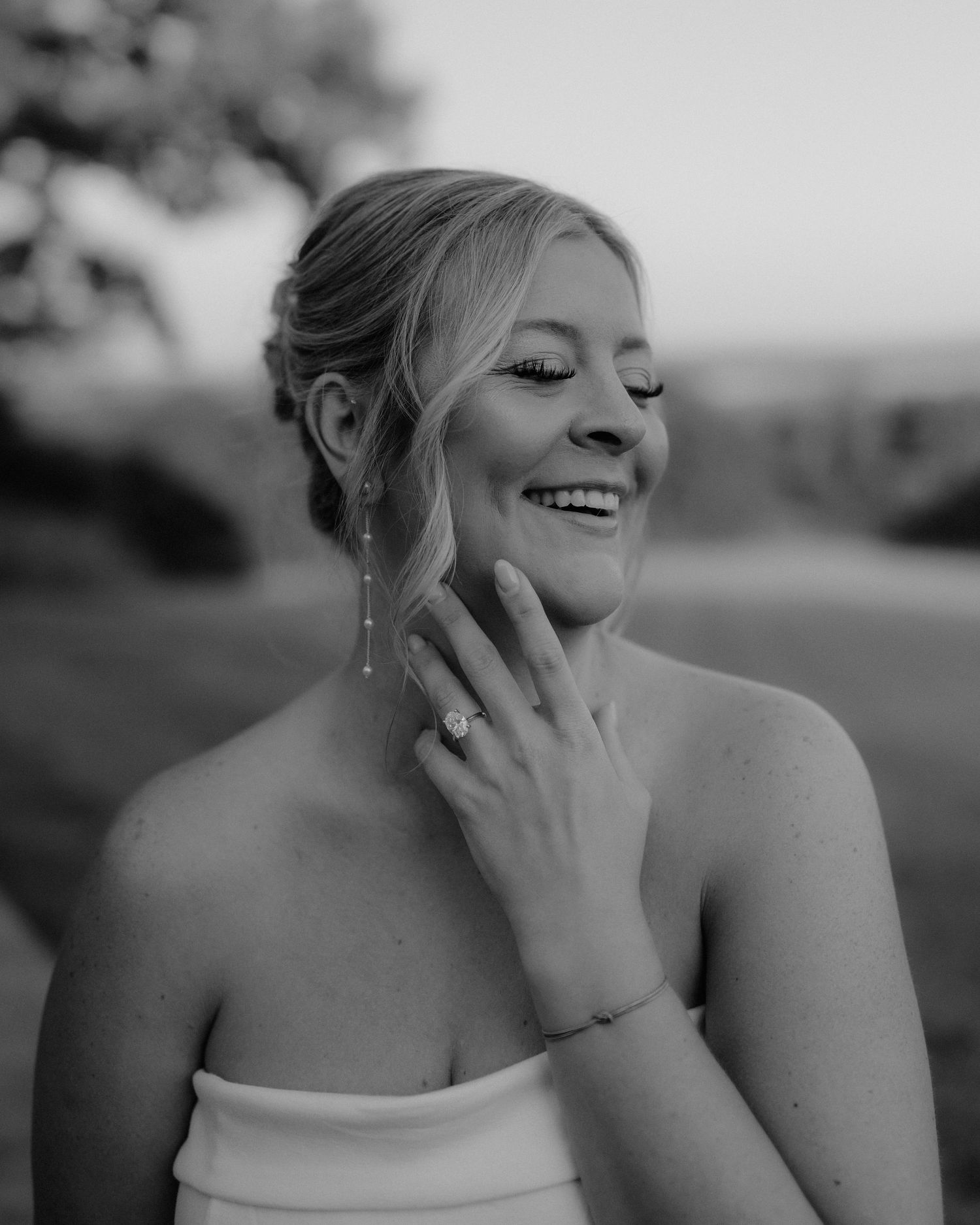 Black and white photo of a smiling bride in strapless gown, wearing drop earrings and engagement ring.