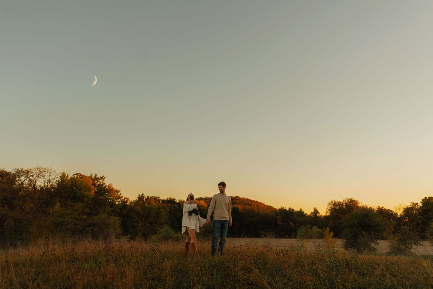Couple holding hands in a golden field at sunset with a crescent moon and trees in the background.