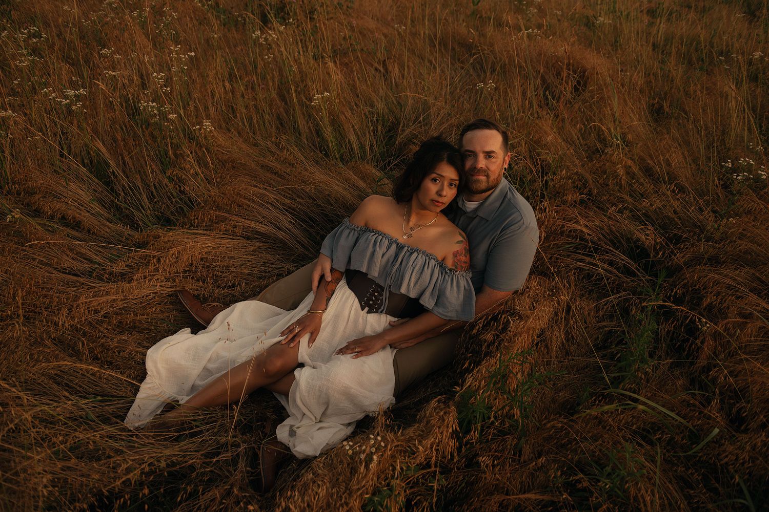 A couple relaxes together in a golden hay field at sunset, woman in flowing white dress.