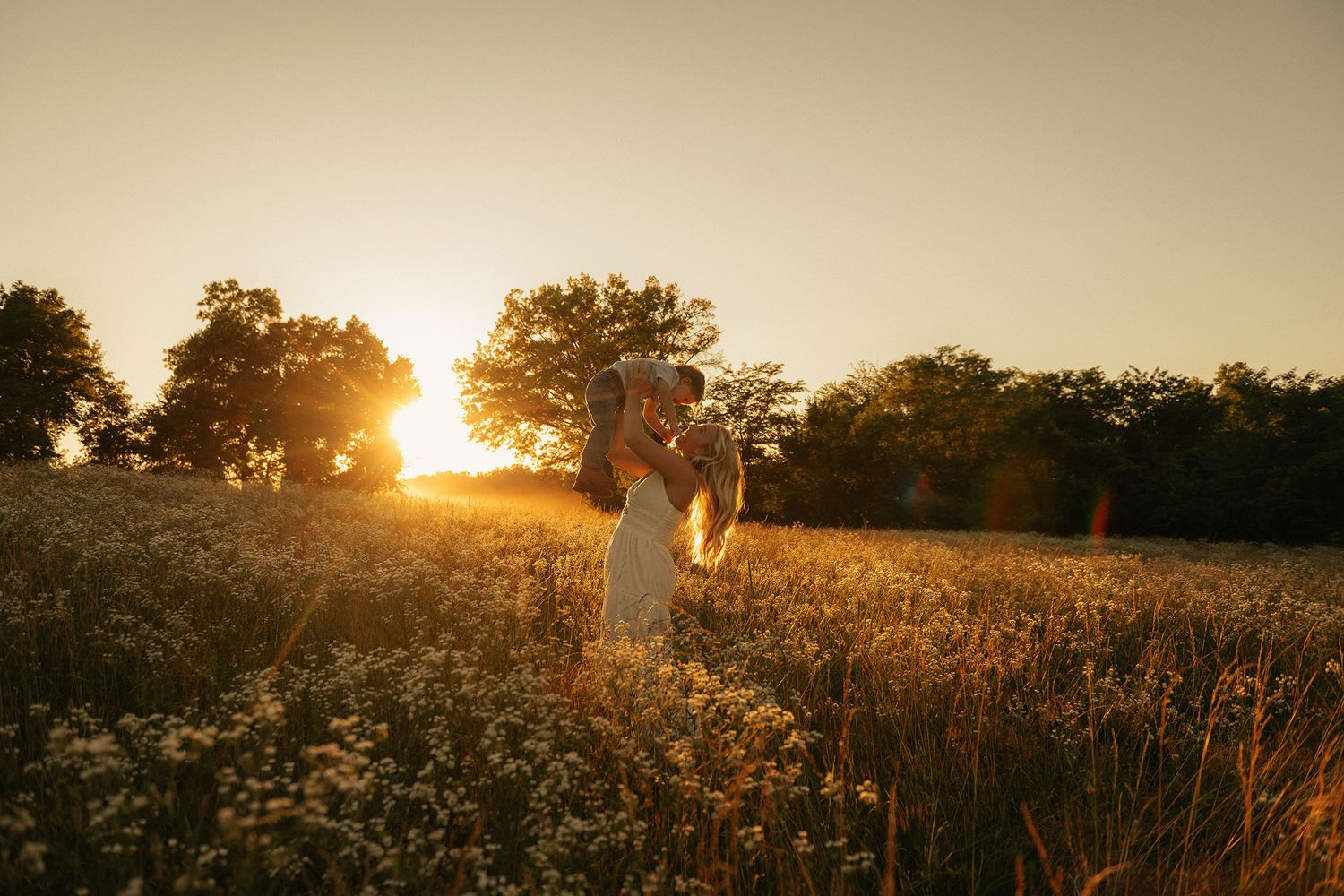 A woman in white holds a child in a wildflower meadow bathed in warm golden sunset light.