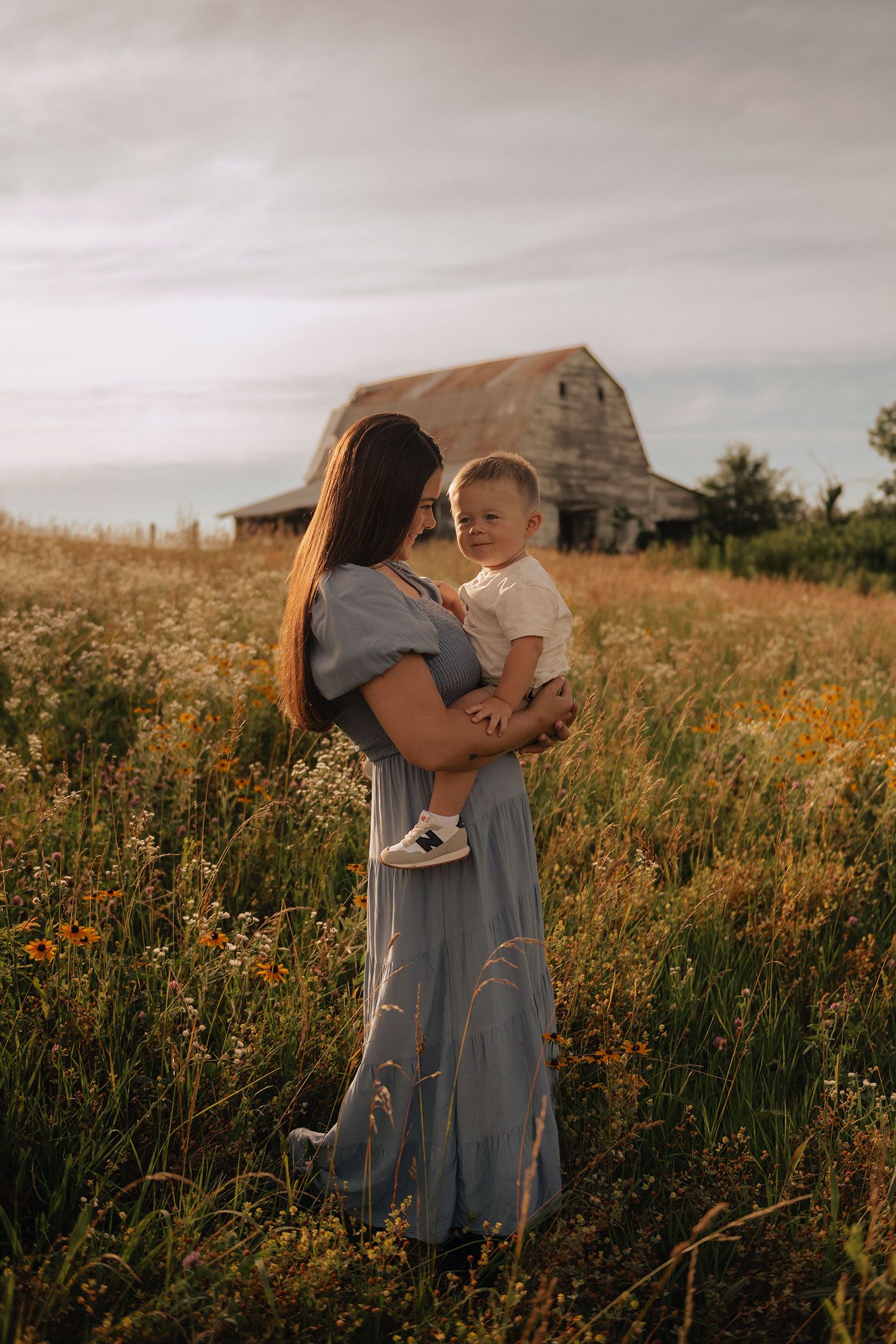 A mother in a blue dress holds her toddler son in a wildflower field near an old barn.