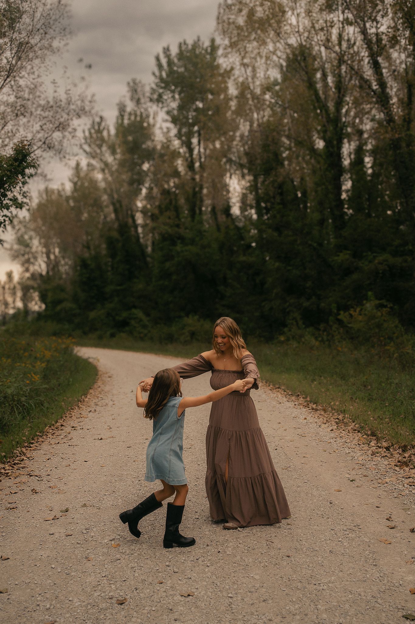 Woman in brown dress dancing with young girl in blue dress on a scenic gravel path surrounded by lush green trees.