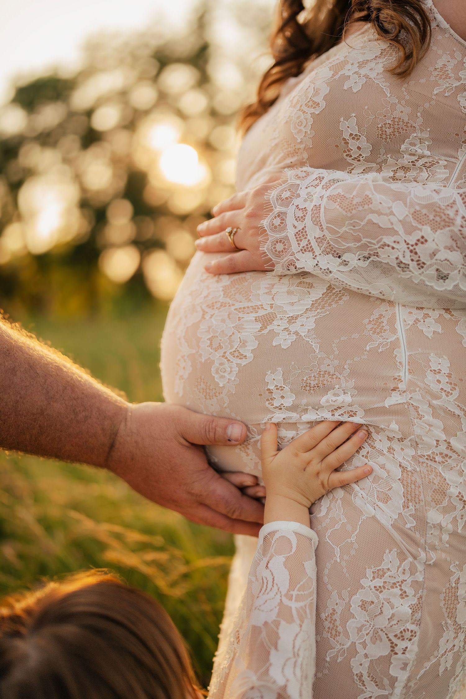 Pregnant woman in white lace dress outdoors at golden hour, with partner and child gently touching her baby bump.