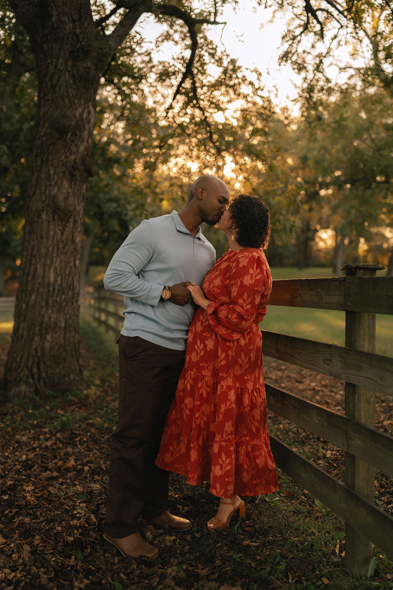 Couple sharing a romantic kiss at golden hour by a wooden fence surrounded by autumn trees.