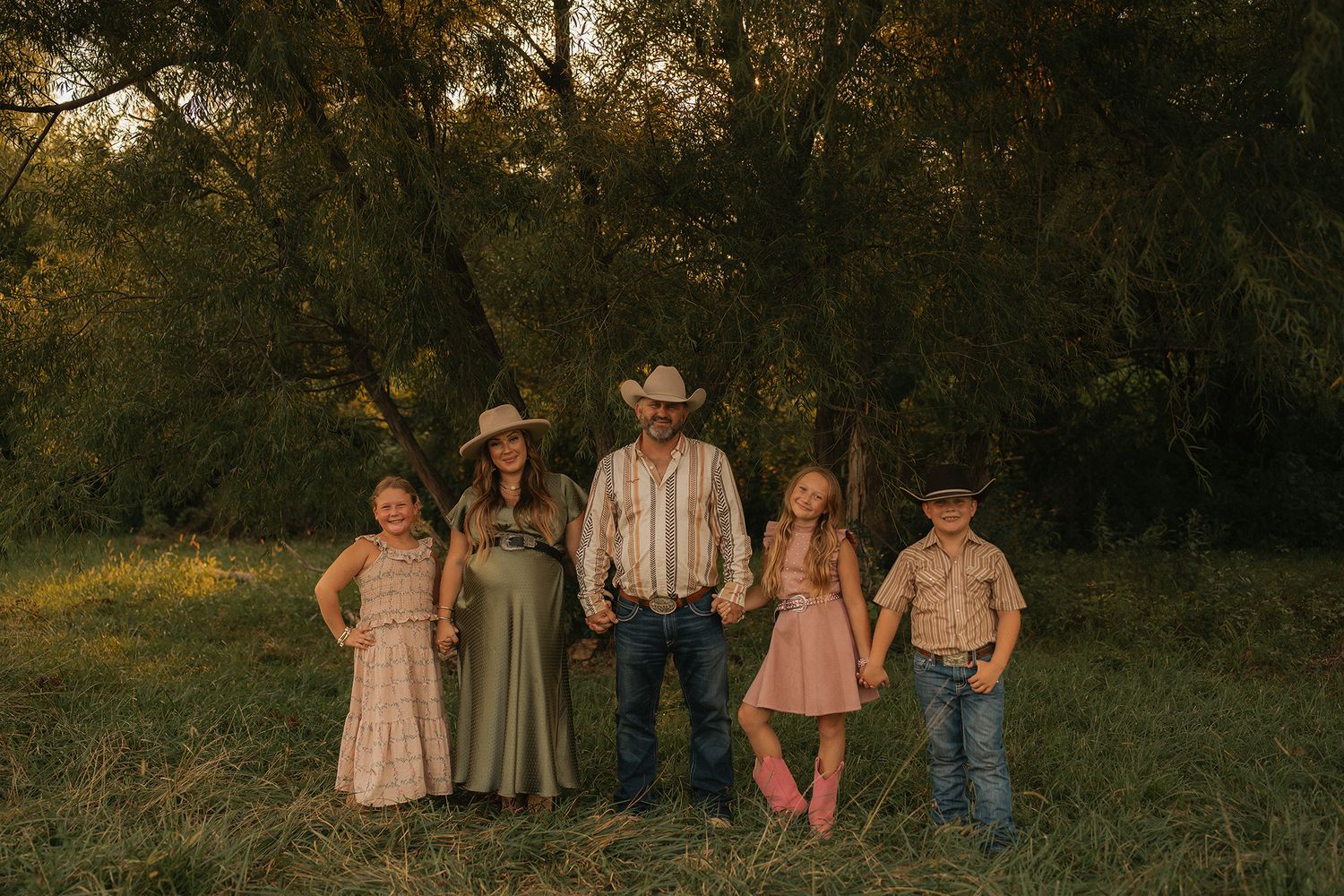 Family of five in cowboy hats posing outdoors in a golden meadow with lush green trees in the background.