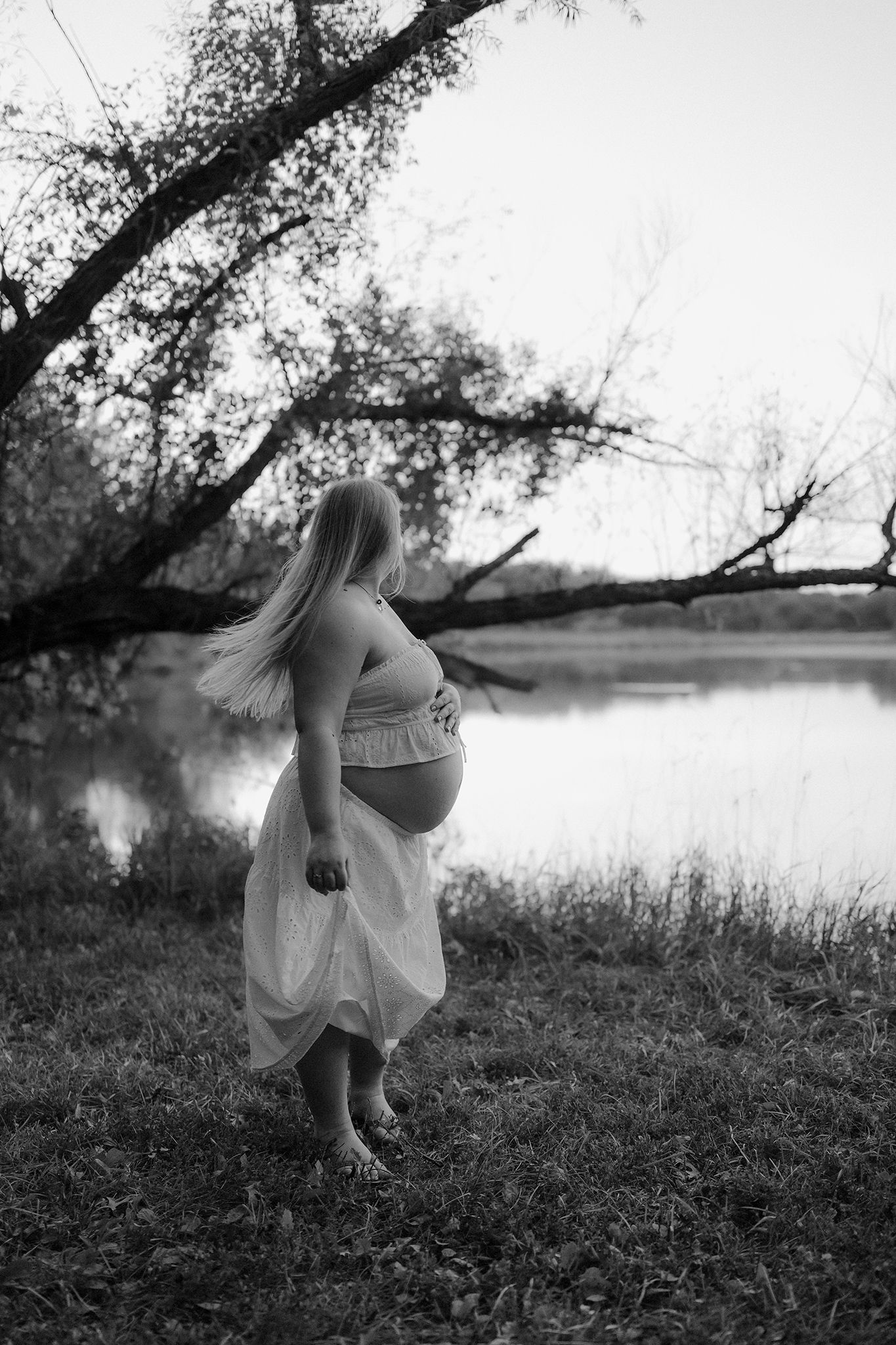 Black and white photo of a pregnant woman standing barefoot by a serene lake, holding her skirt while showing her baby bump.