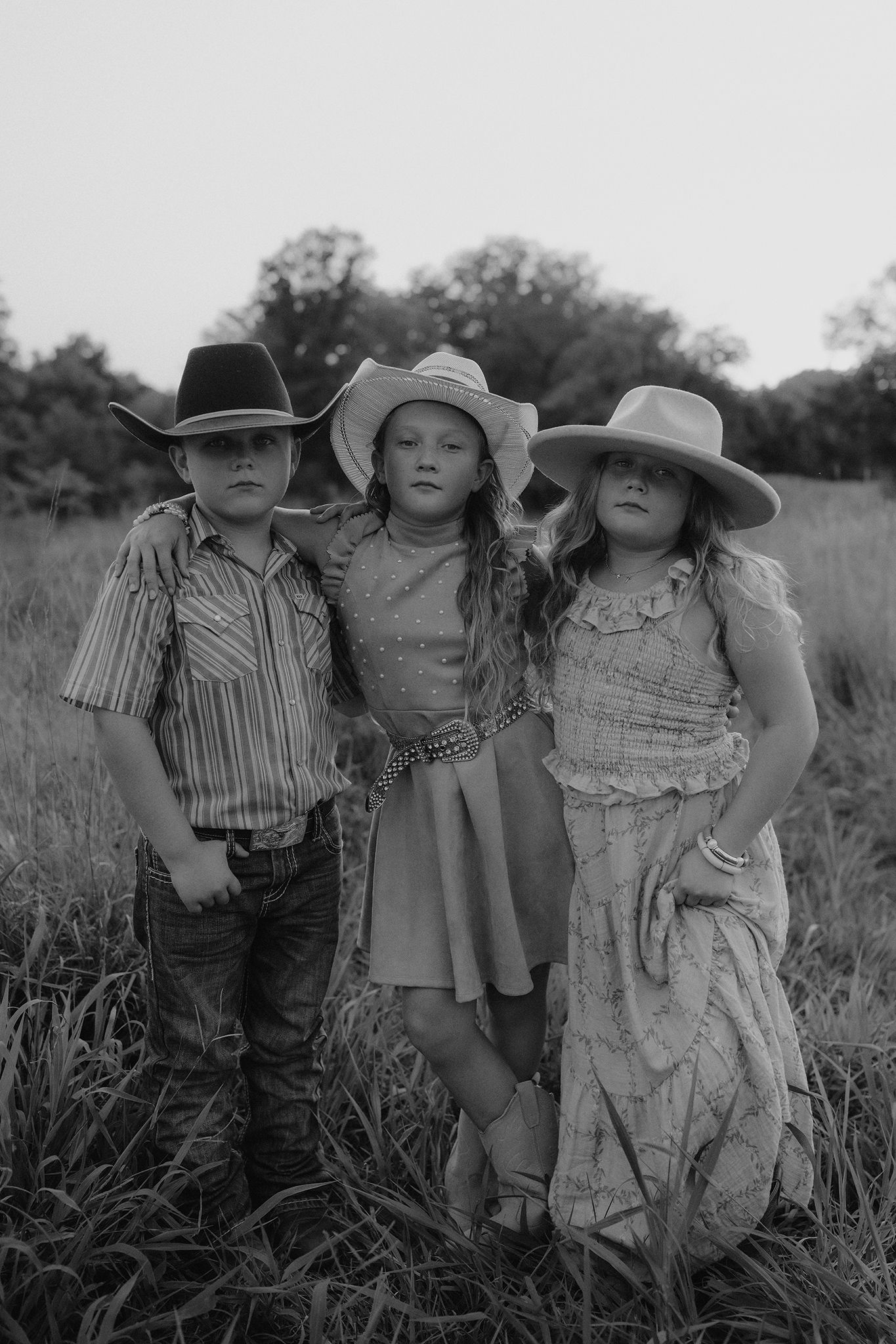 Three children in cowboy hats pose in a field, dressed in western attire, captured in black and white photography.